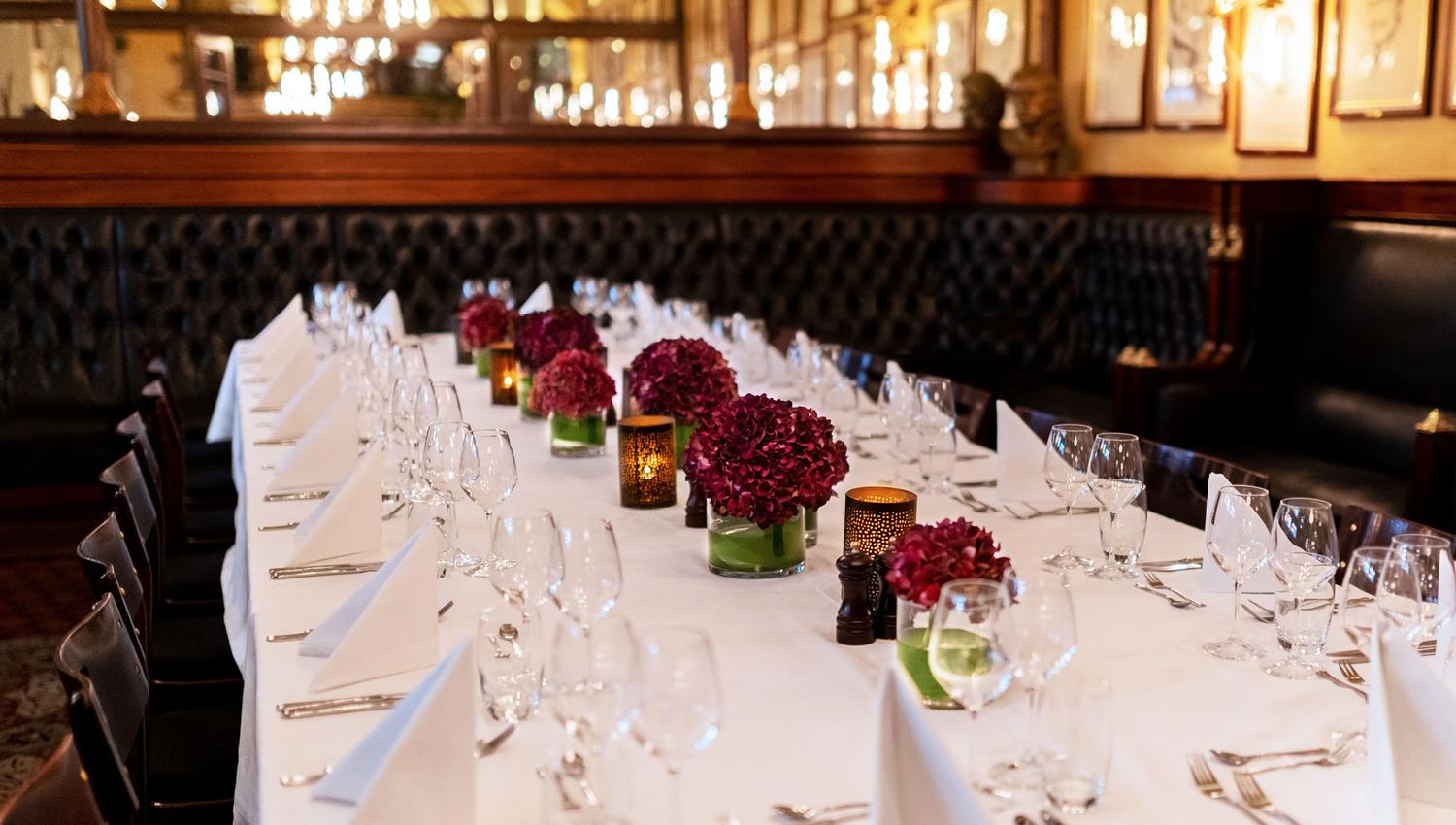 A long table with a white tablecloth and red flowers on, set for dining.