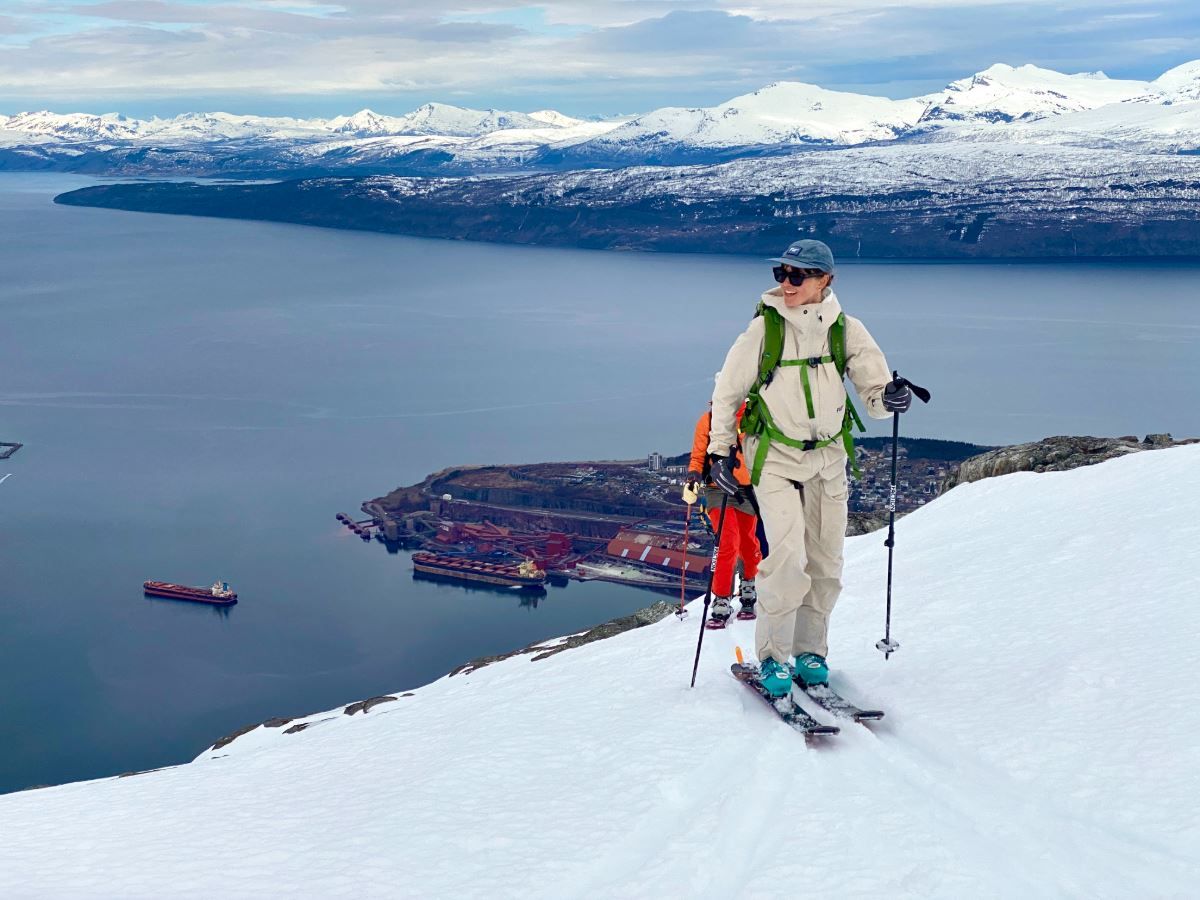 Two skiers climb a snowy slope with poles, overlooking a fjord, harbor below, and distant snowcapped mountains..