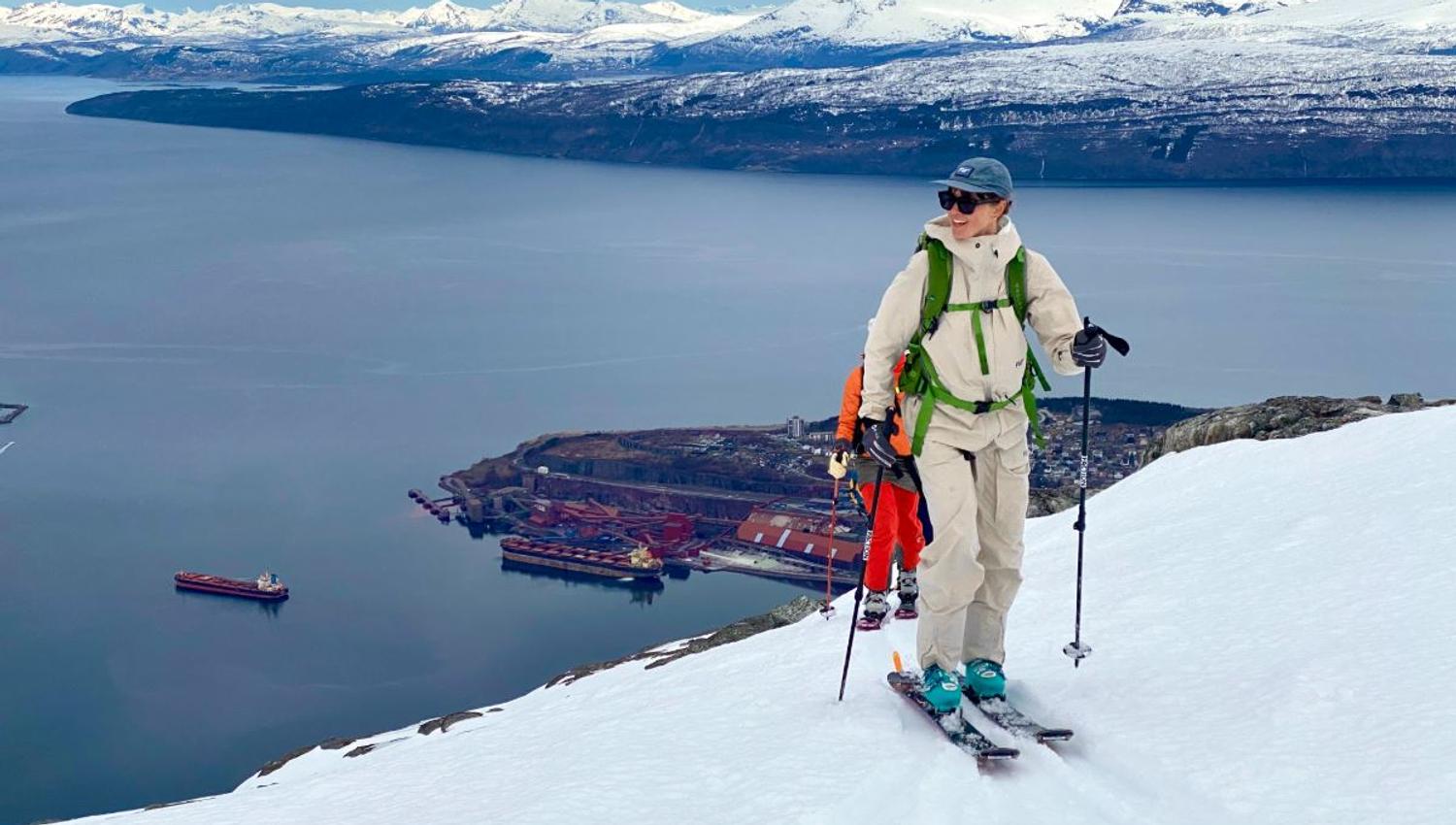 Two skiers climb a snowy slope with poles, overlooking a fjord, harbor below, and distant snowcapped mountains..