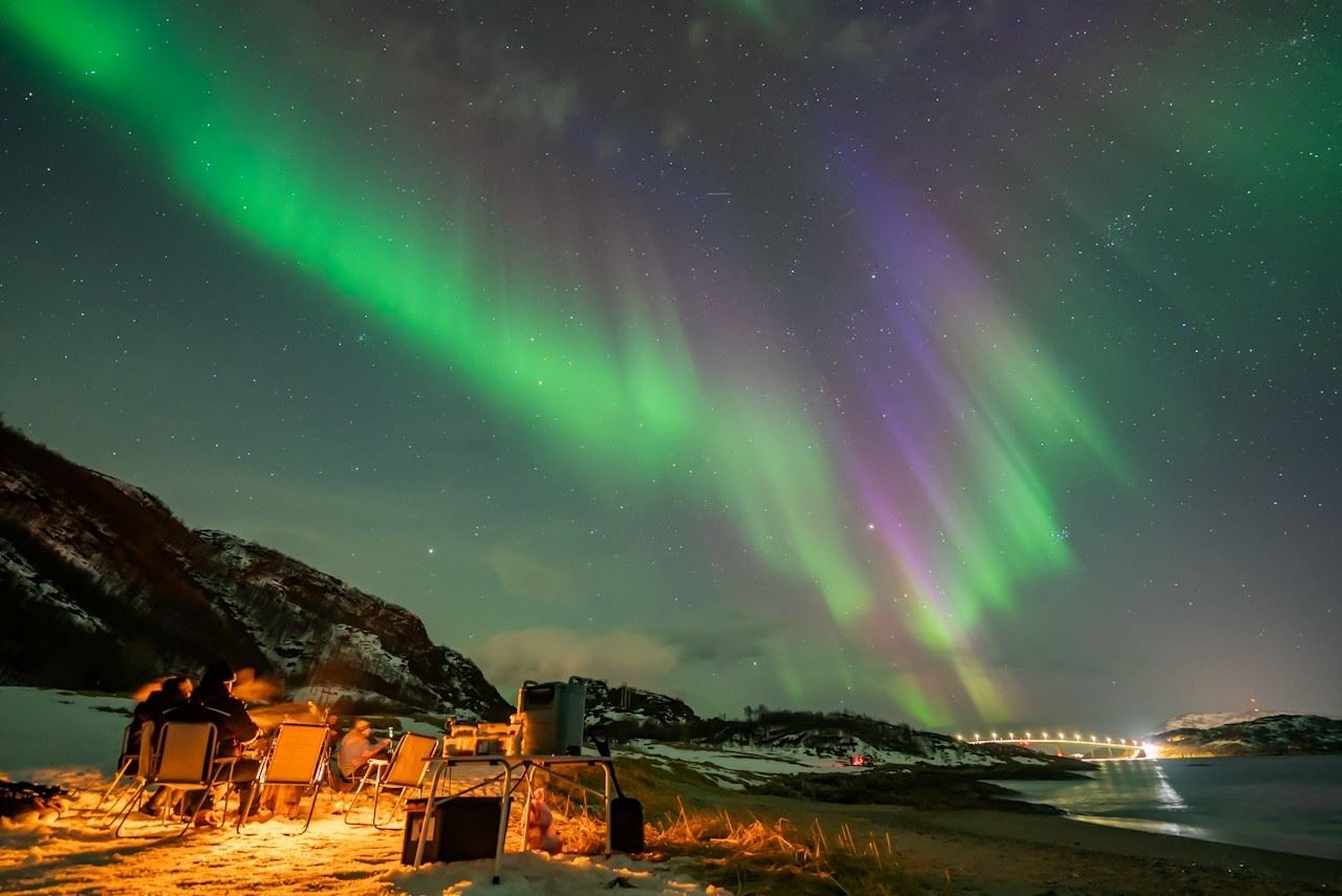 Guests gather around a campfire as the Northern Lights appear in the sky during a Northern Lights chase.