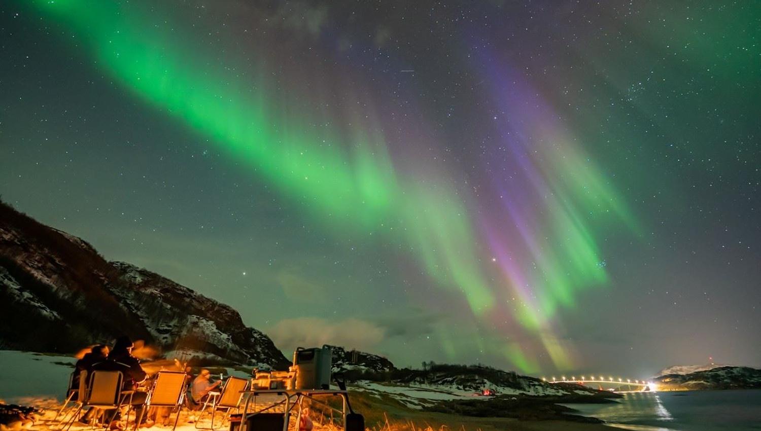 Guests gather around a campfire as the Northern Lights appear in the sky during a Northern Lights chase.
