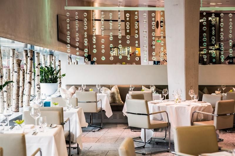 Restaurant interior with a range of white, clothed tables, beige leather chairs, beige walls, a pillar, and a decoration hanging from the ceiling.