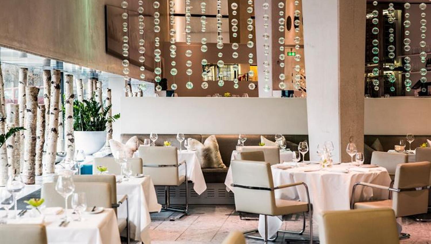Restaurant interior with a range of white, clothed tables, beige leather chairs, beige walls, a pillar, and a decoration hanging from the ceiling.