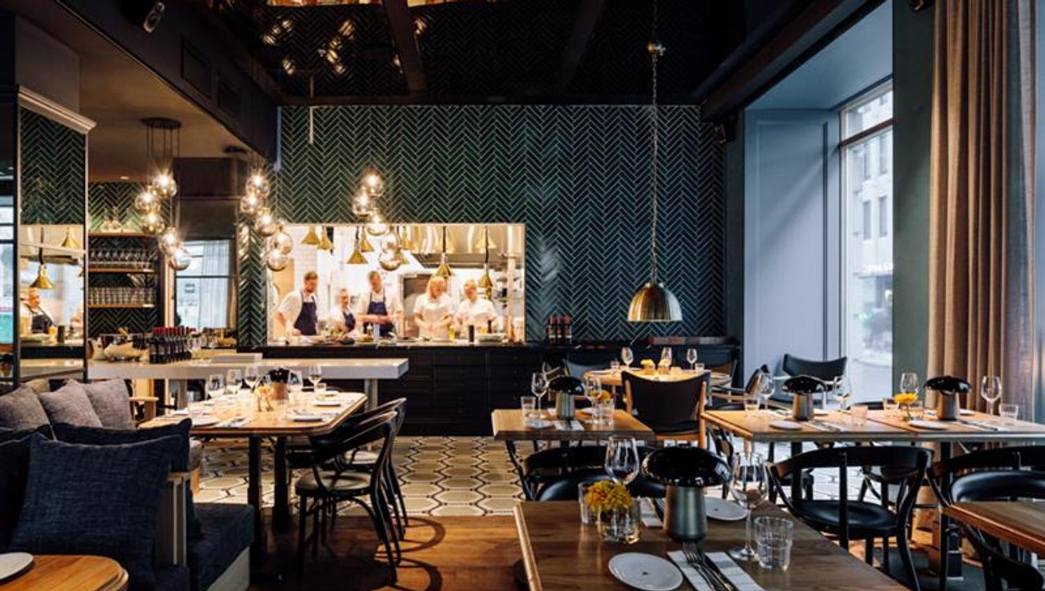 Restaurant Atlas - interiørbilde. Dekkede bord med stoler, sitteseksjon, hengende taklamper og kjøkken i bakgrunnen.