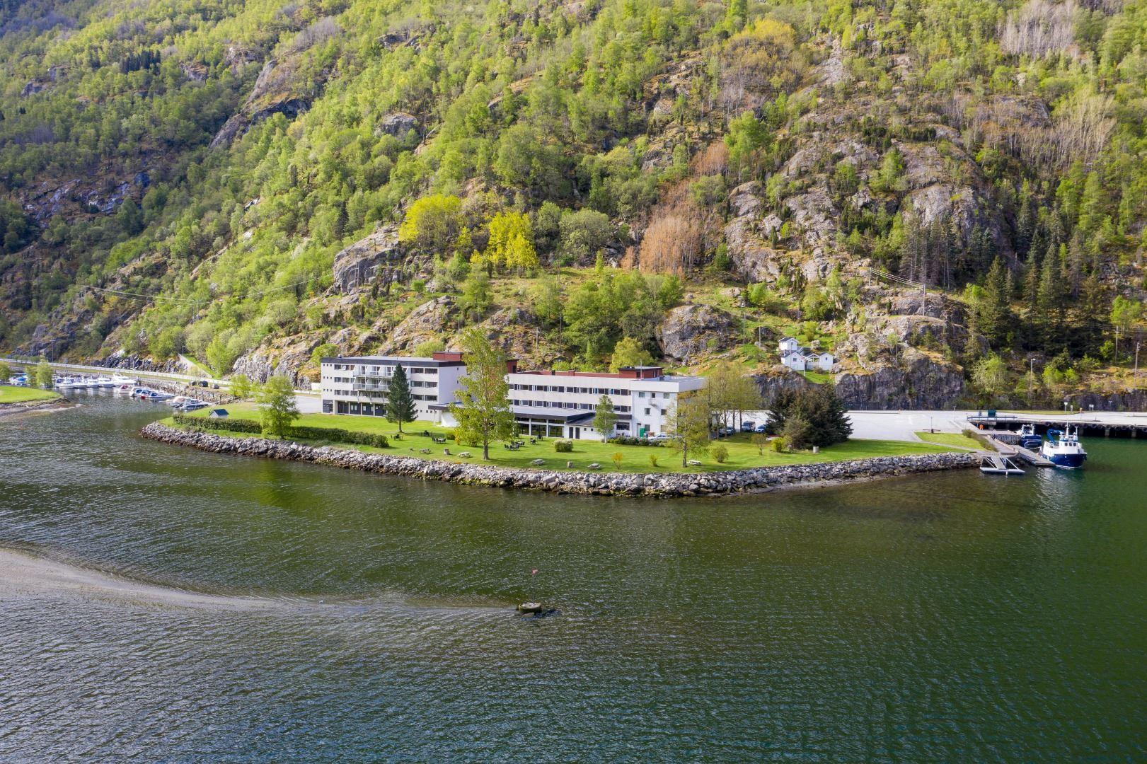 A long white building on a grassy shoreline beside a calm lake, backed by steep rocky hills covered in trees..