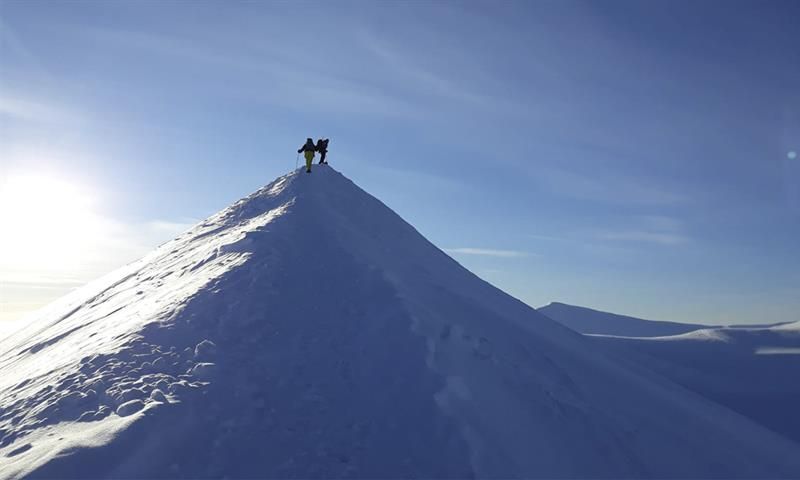 Two persons on top of a snowy mountain ridge with snow-covered landscapes and a blue sky in the background