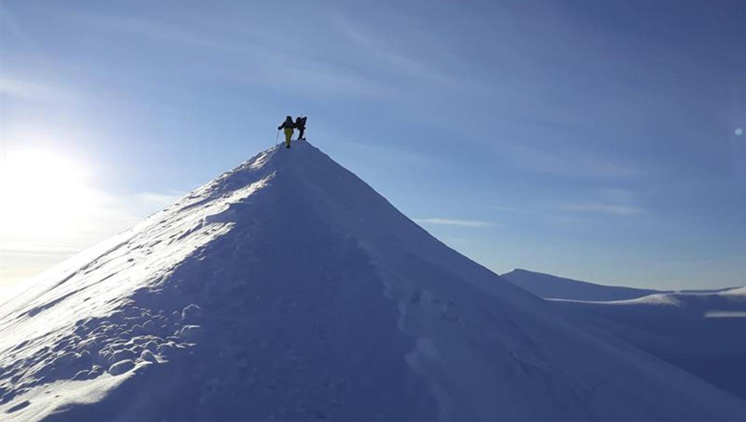Two persons on top of a snowy mountain ridge with snow-covered landscapes and a blue sky in the background