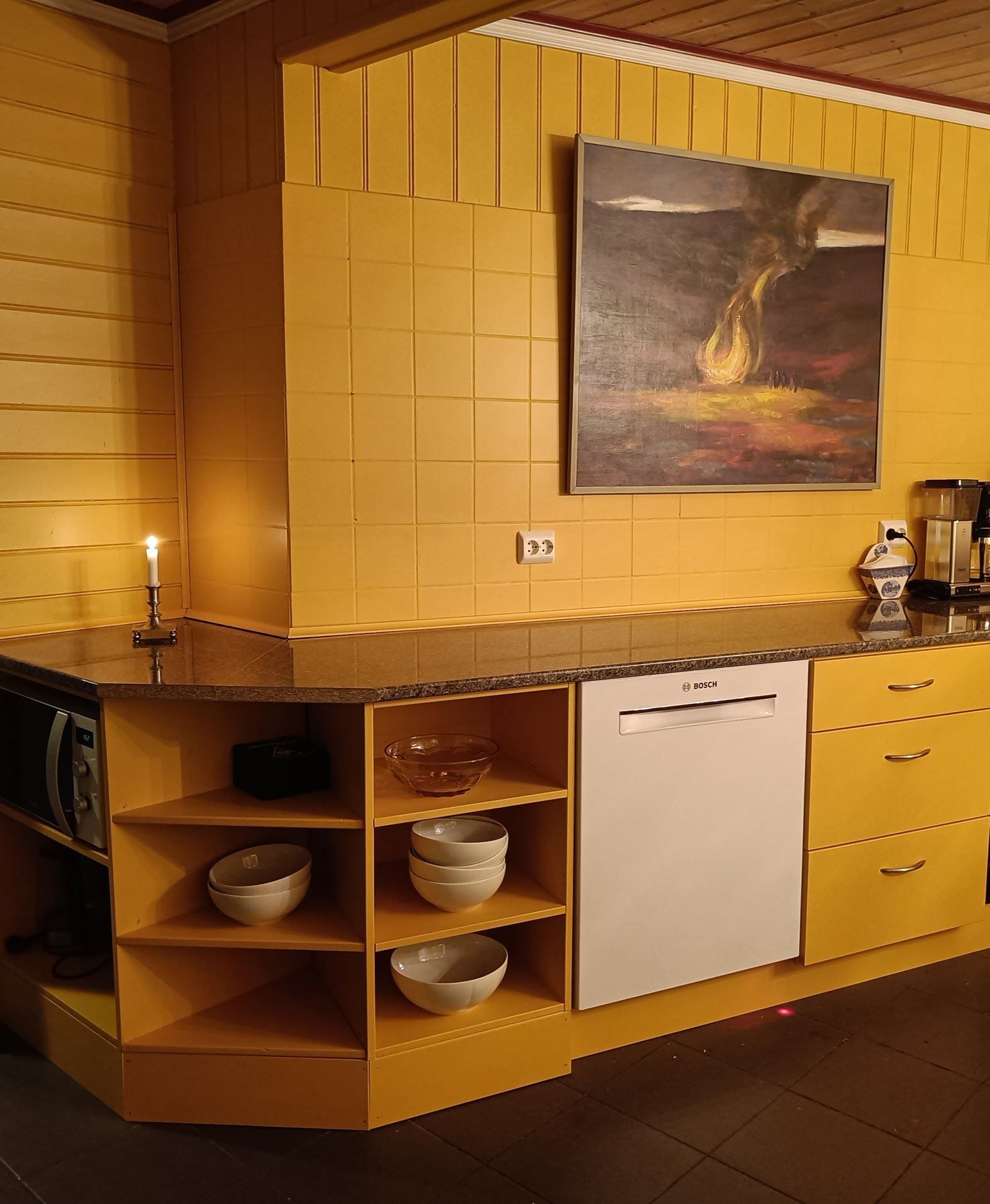 Orheimstunet in Nordfjord - Yellow-tiled kitchen counter with open shelves of bowls, a dishwasher, candle, coffee maker, and a framed painting..