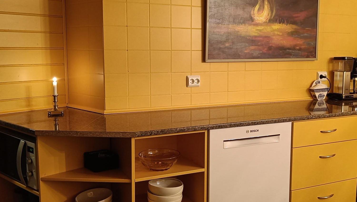 Orheimstunet in Nordfjord - Yellow-tiled kitchen counter with open shelves of bowls, a dishwasher, candle, coffee maker, and a framed painting..