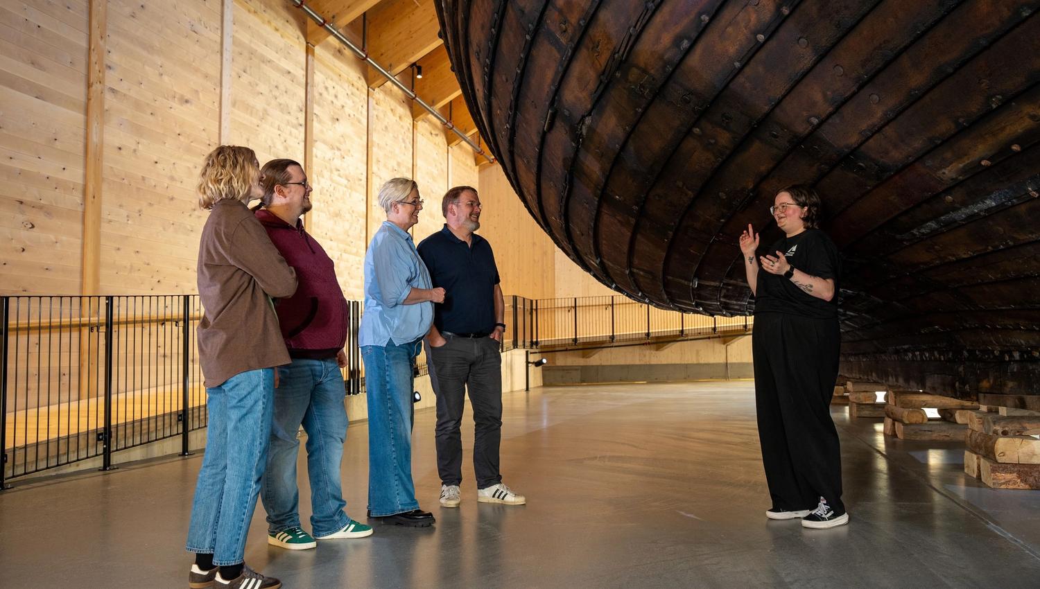 A small group listens to a guide inside a wooden hall beneath a large suspended wooden ship hull..