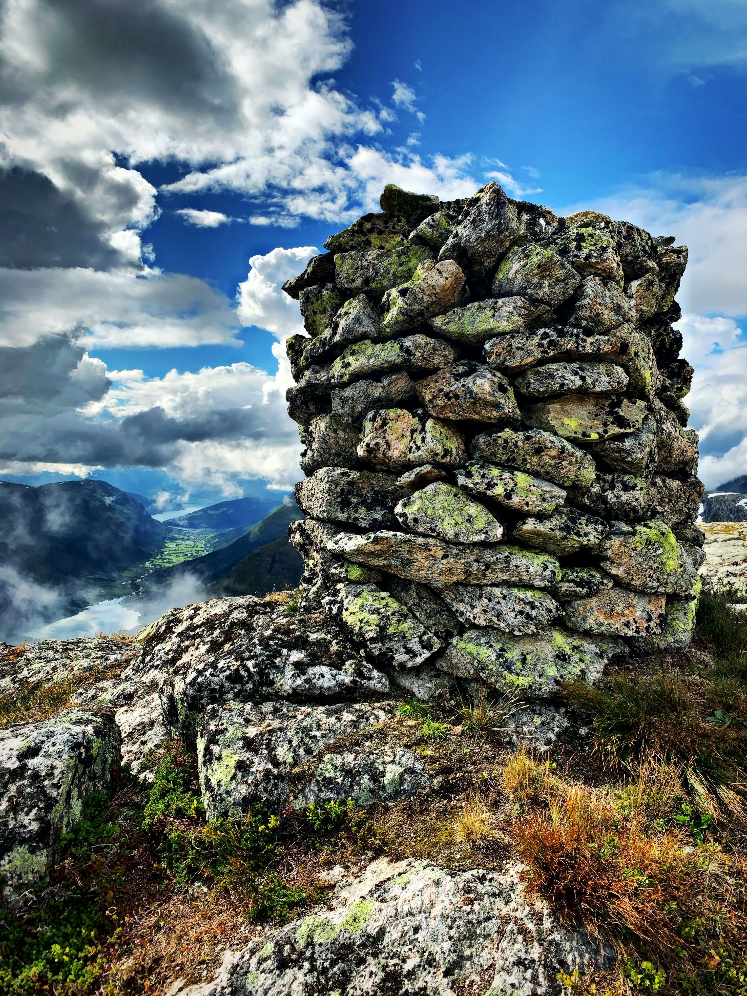 Mossy stacked-stone cairn on a rocky mountaintop, with valley and lake below under dramatic clouds and blue sky..