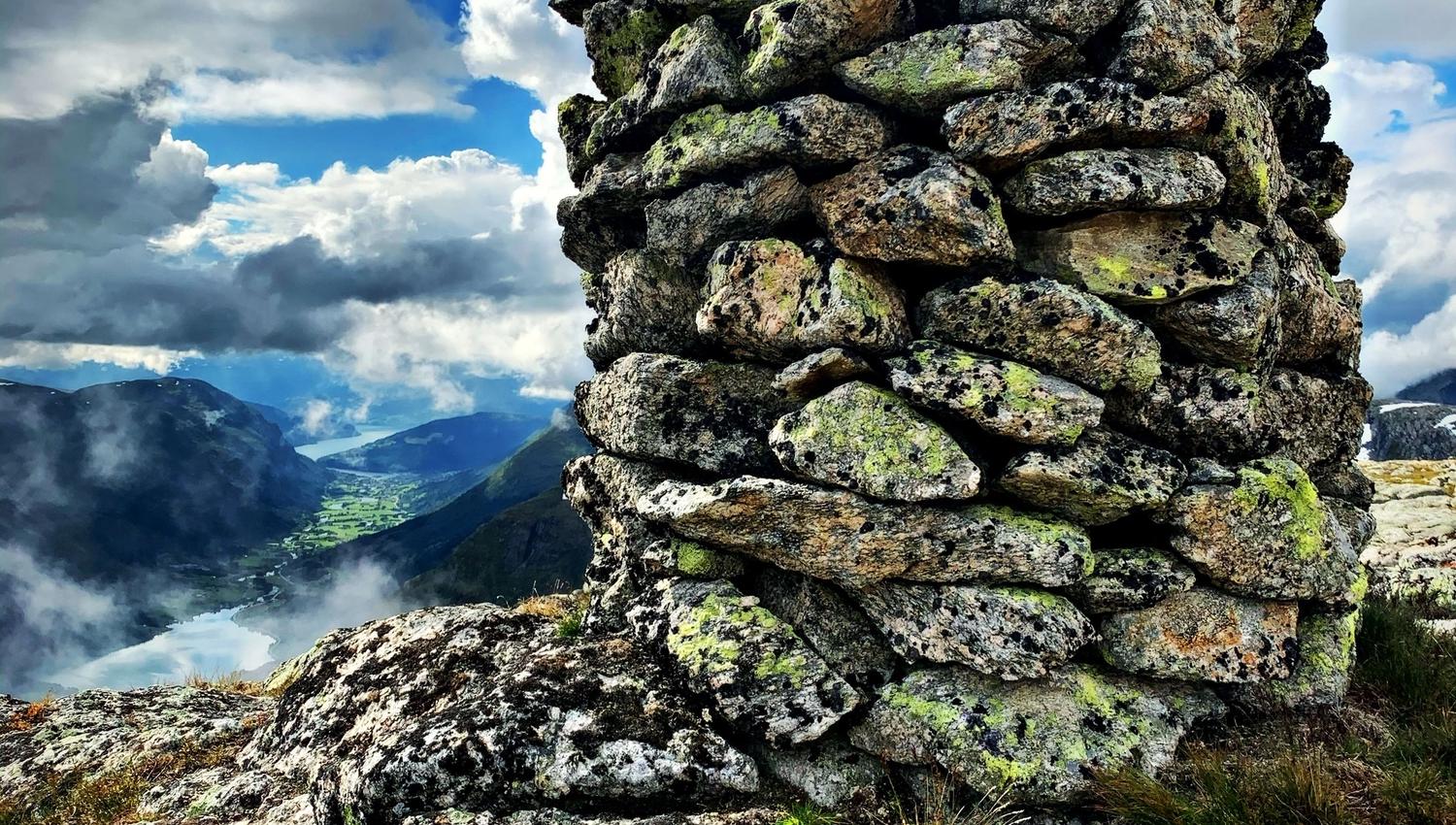 Mossy stacked-stone cairn on a rocky mountaintop, with valley and lake below under dramatic clouds and blue sky..