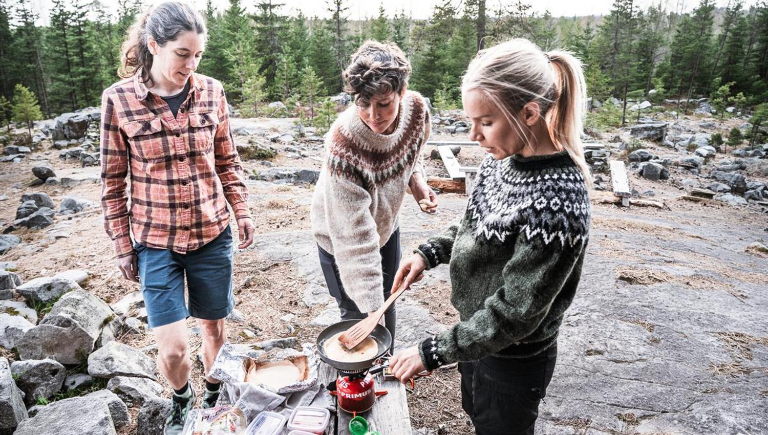 Three people cooking outdoors by a fire area.