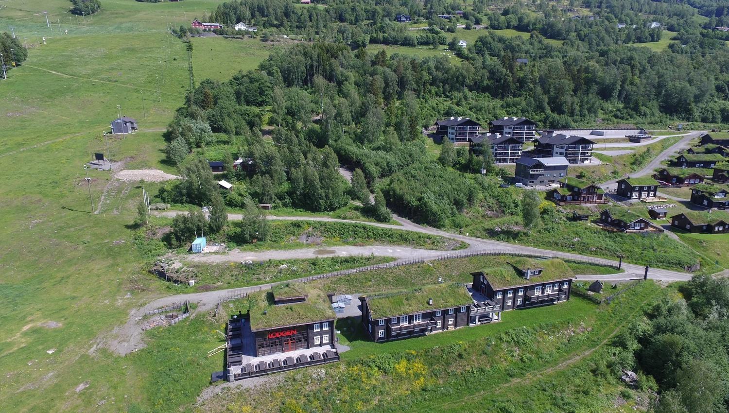 Flygvy över grön fjällsluttning med skog, vägar och flera lodger och stugor under blå himmel.