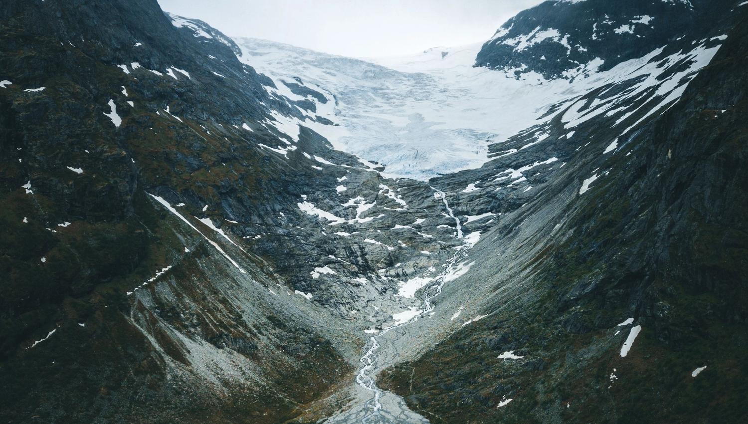 A rugged mountain valley with patches of snow, a glacier at the head, and a pale blue meltwater lake below.