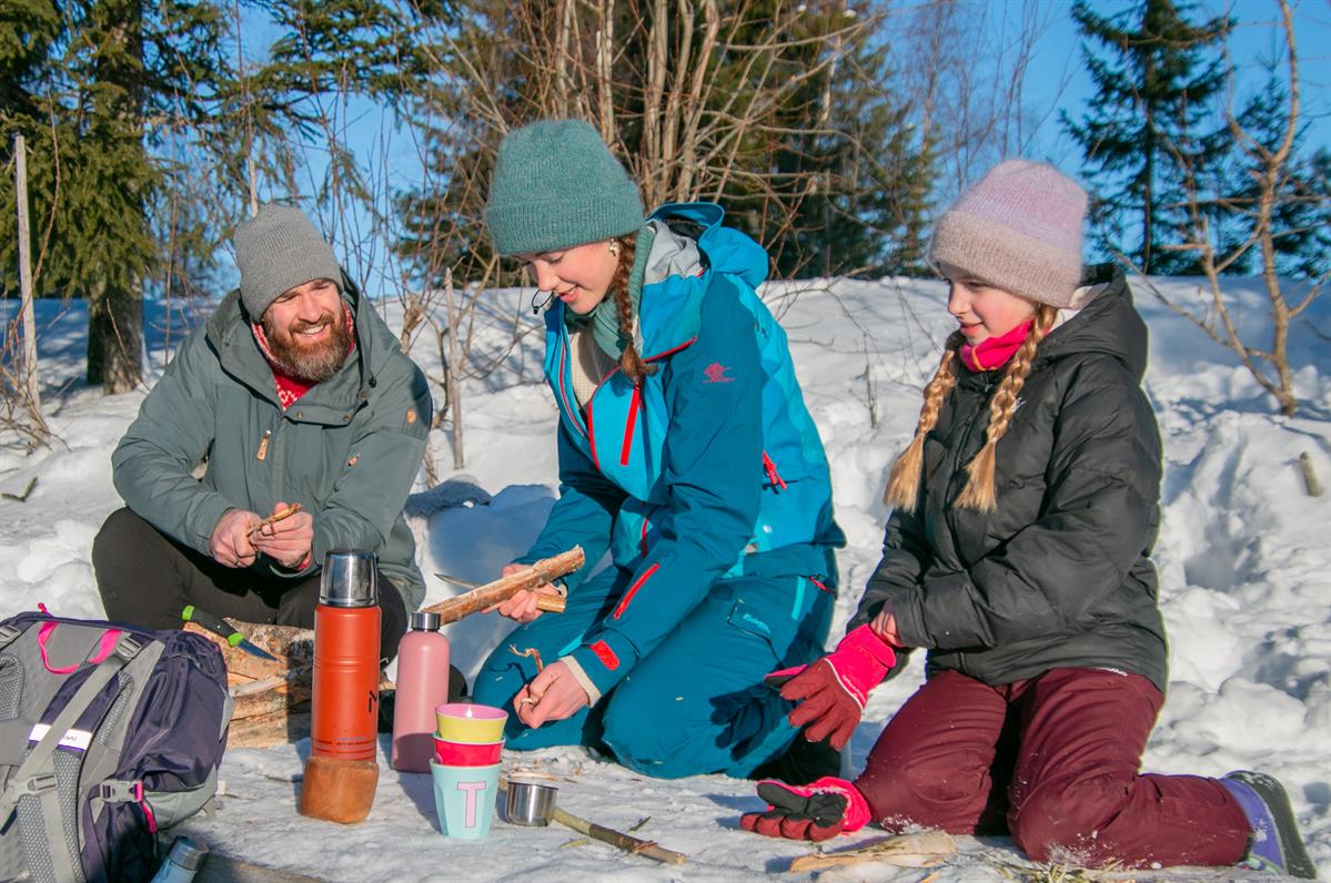 Familie sitter i snøen og deler mat og varm drikke på en vinterdag.