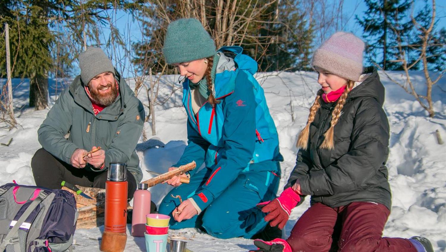 Familie sitter i snøen og deler mat og varm drikke på en vinterdag.