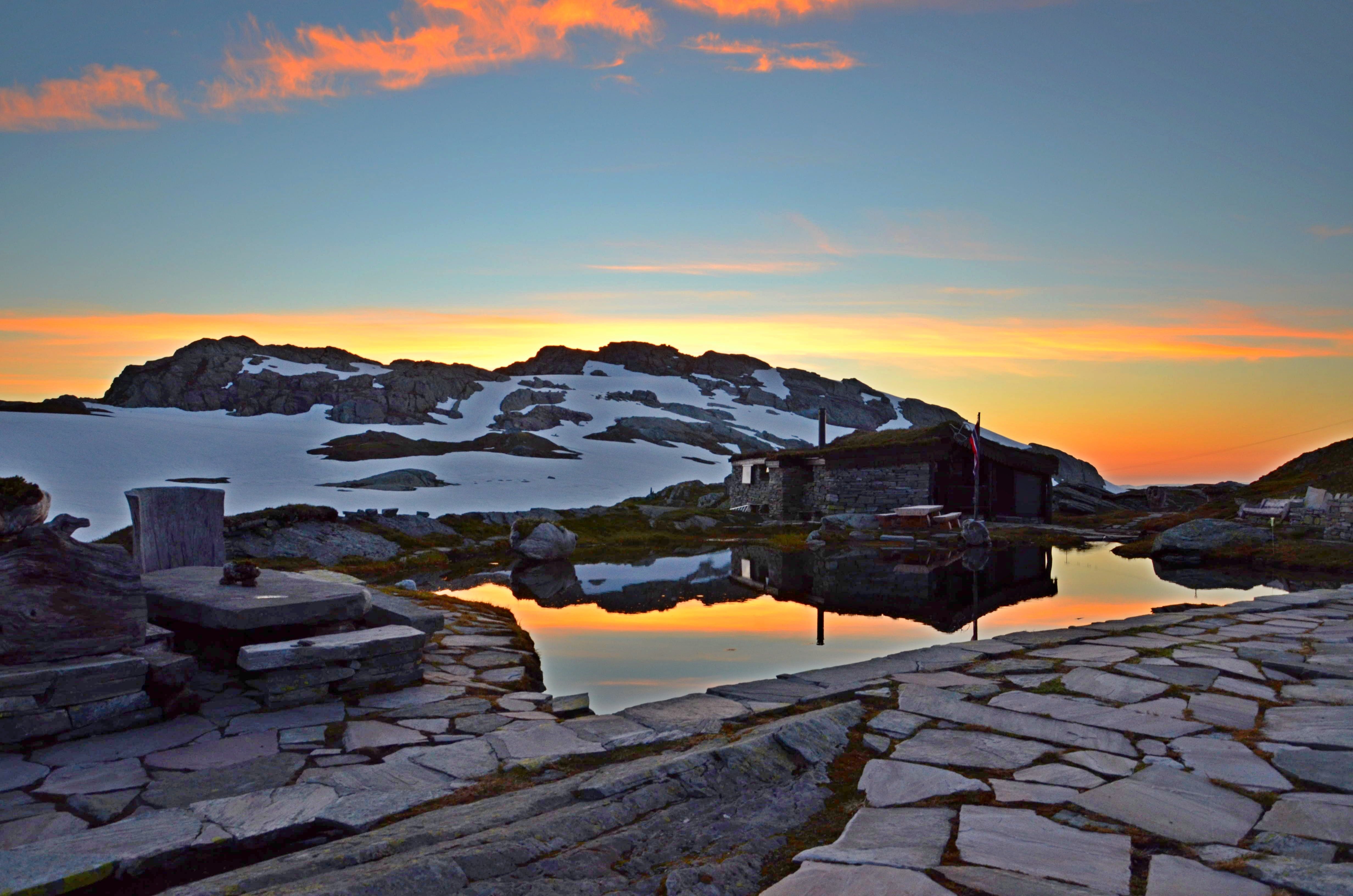 Sunset at Breidablik with a calm mountain pond, stone pathway, and snow-covered peaks in the distance.