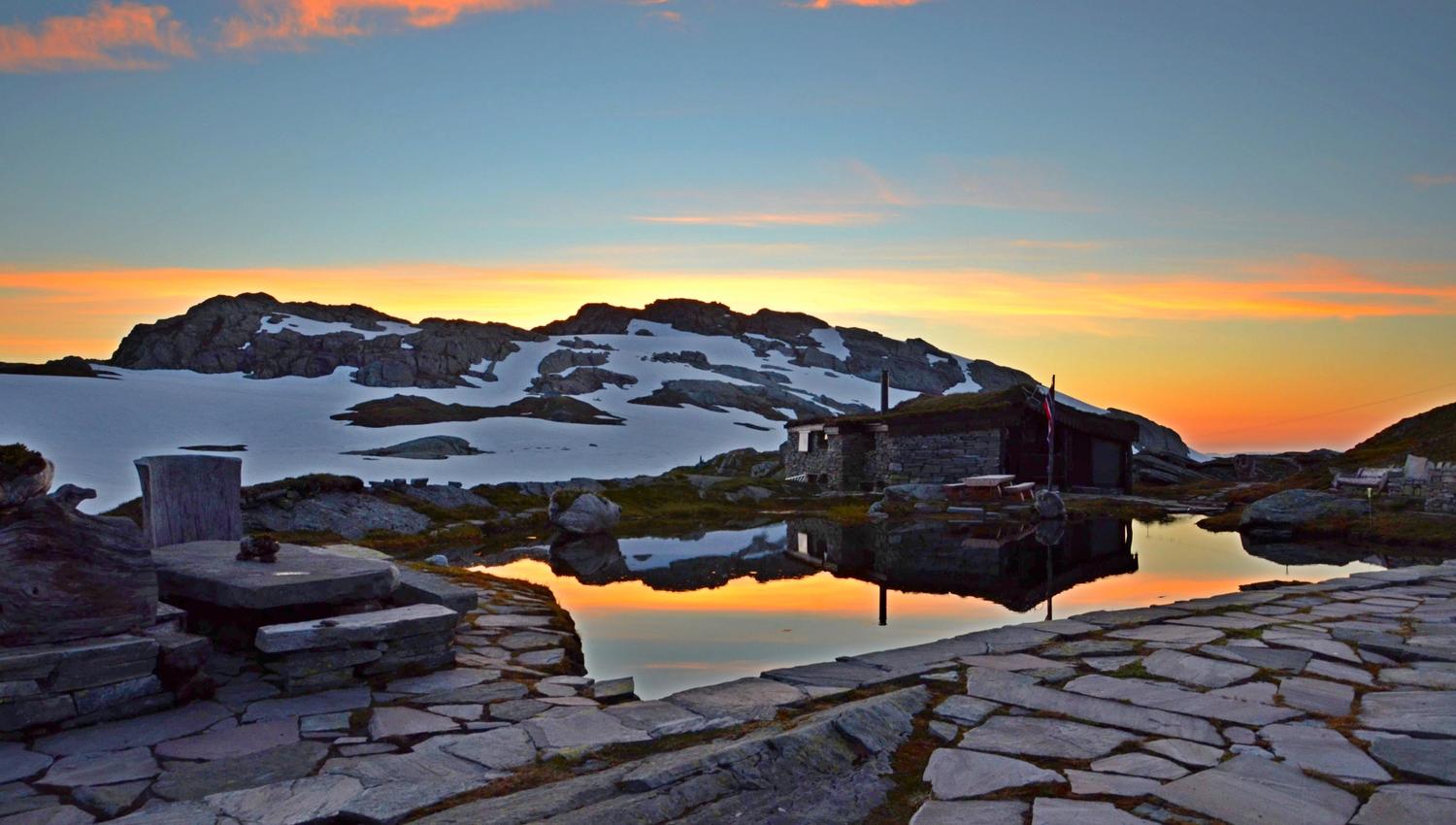 Sunset at Breidablik with a calm mountain pond, stone pathway, and snow-covered peaks in the distance.