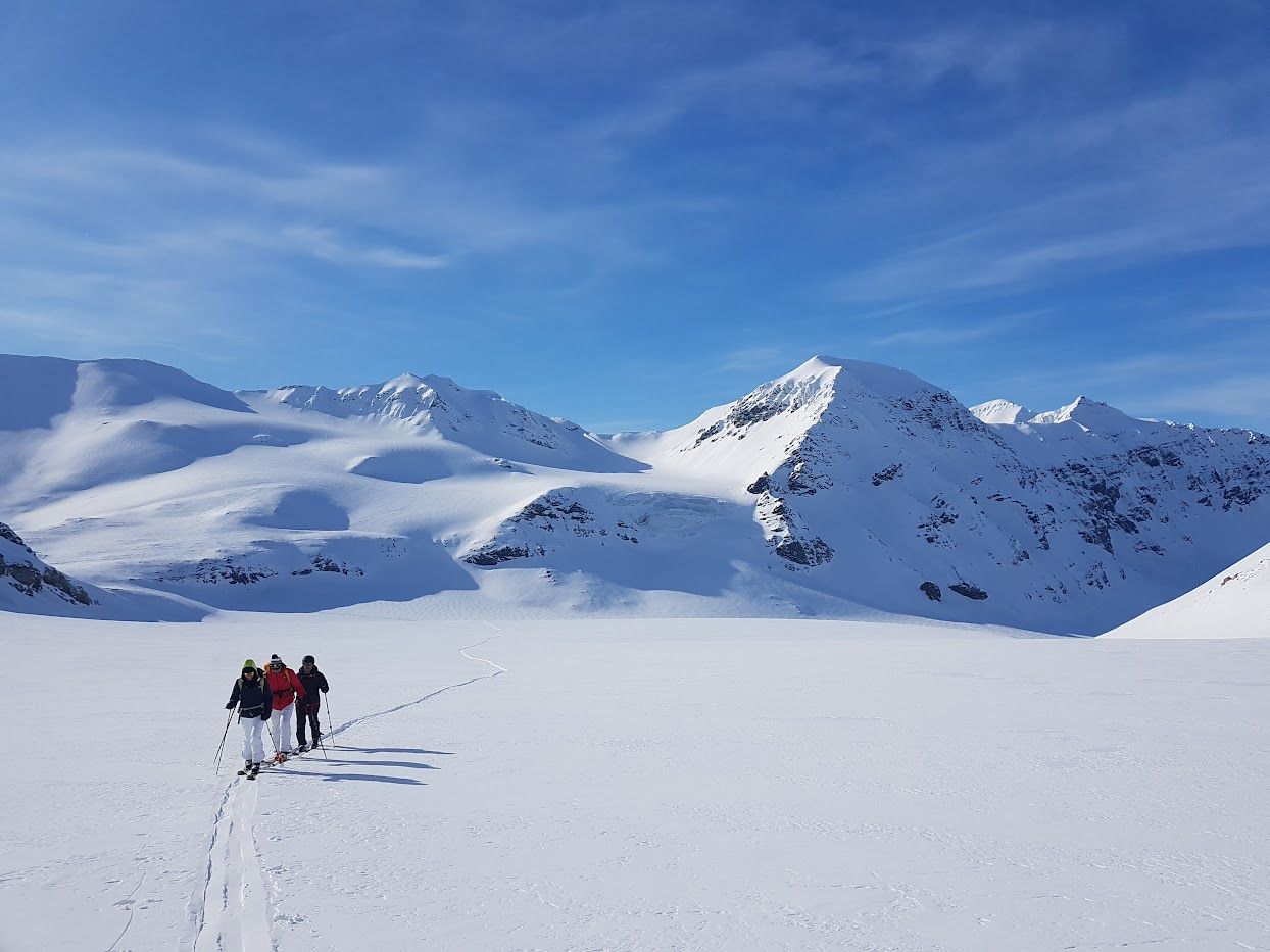Three persons skiing in a line across a flat plateau with tall mountains in the background