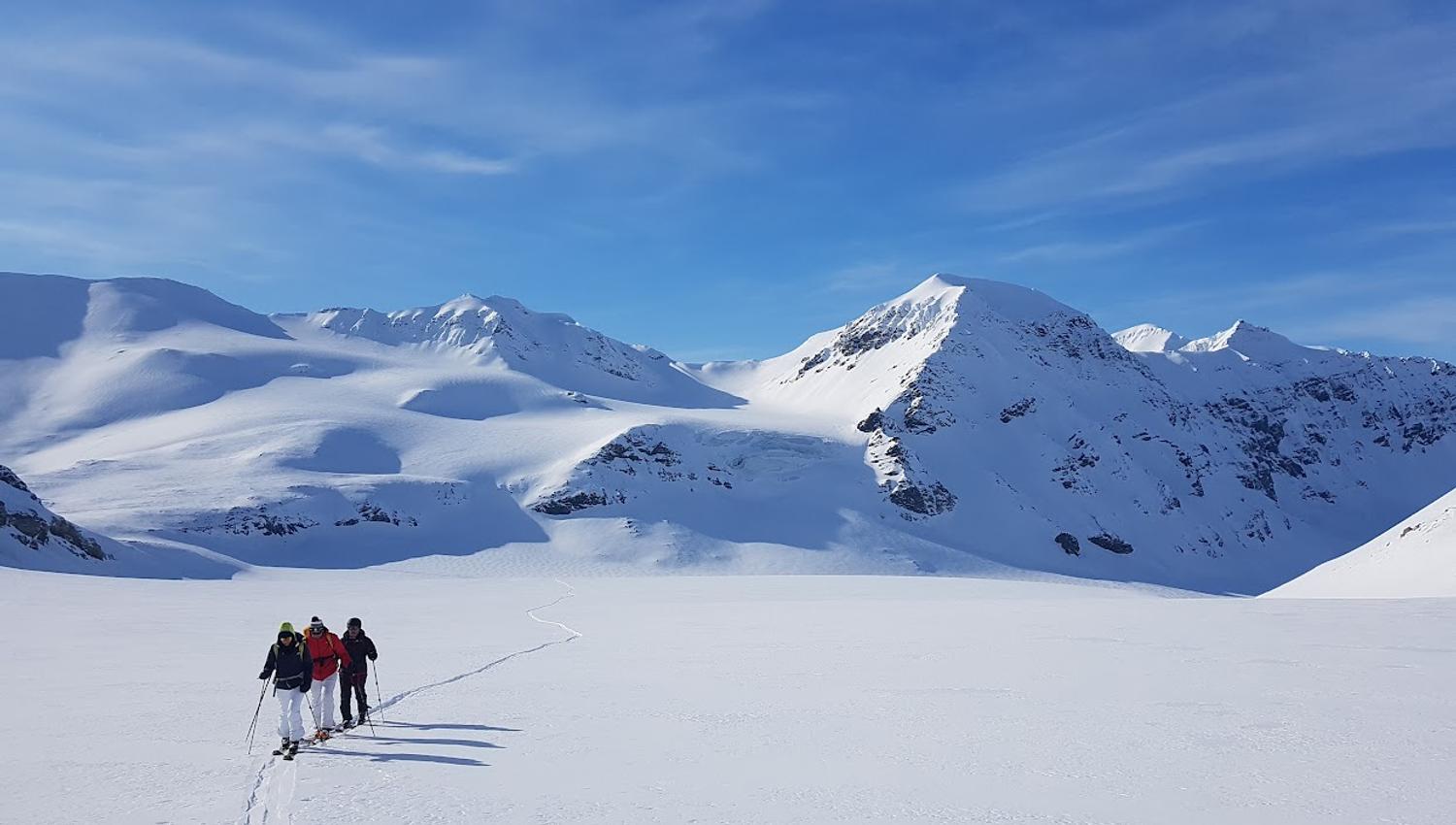 Three persons skiing in a line across a flat plateau with tall mountains in the background