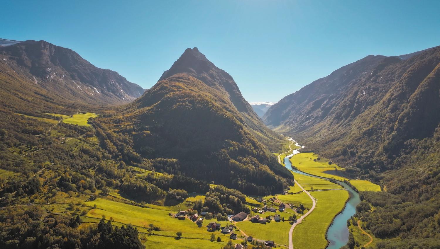 Aerial view of a green valley with a winding river, small village, and steep mountains under a clear blue sky..