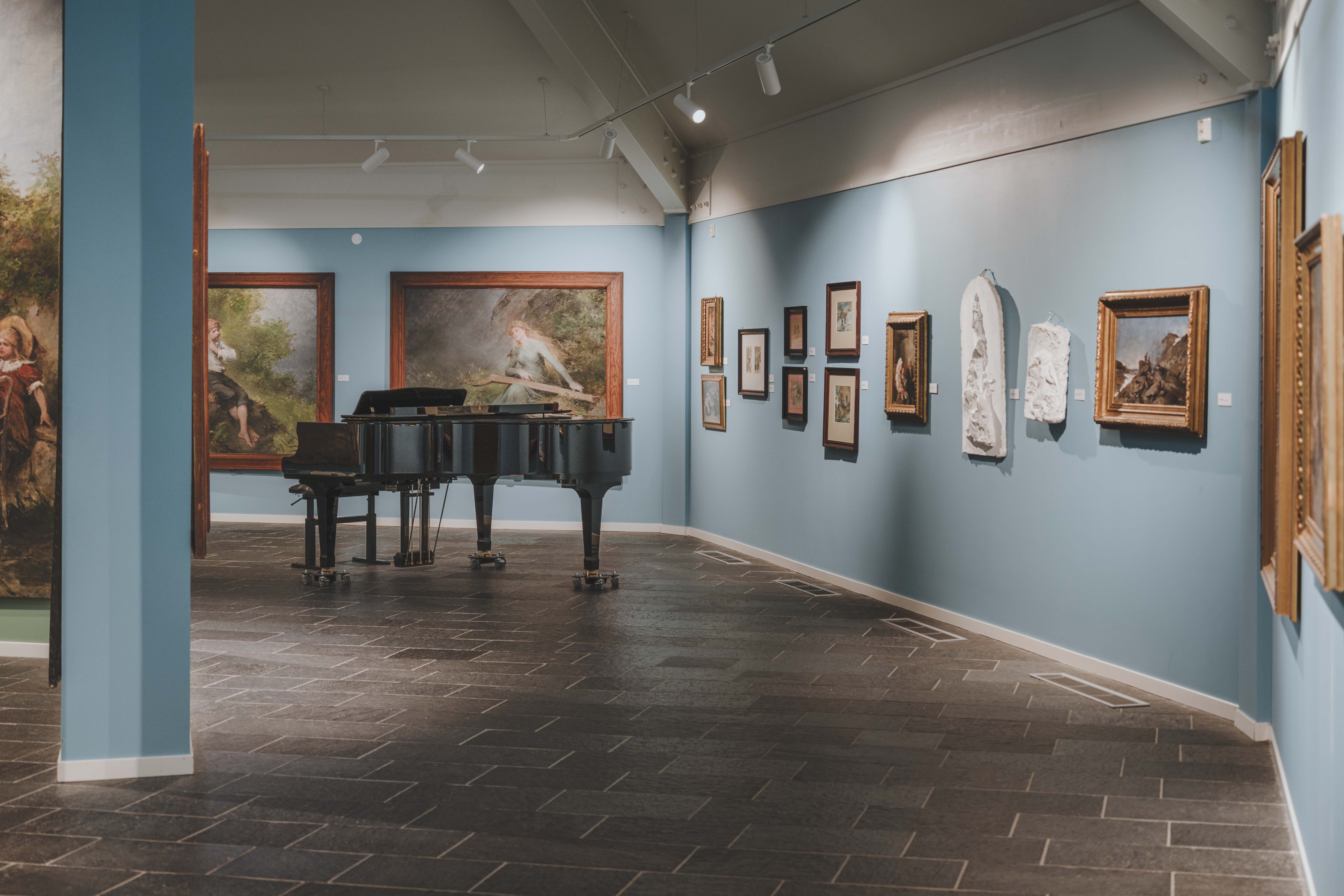 A baby grand piano in front of some of Bergsliens paintings.