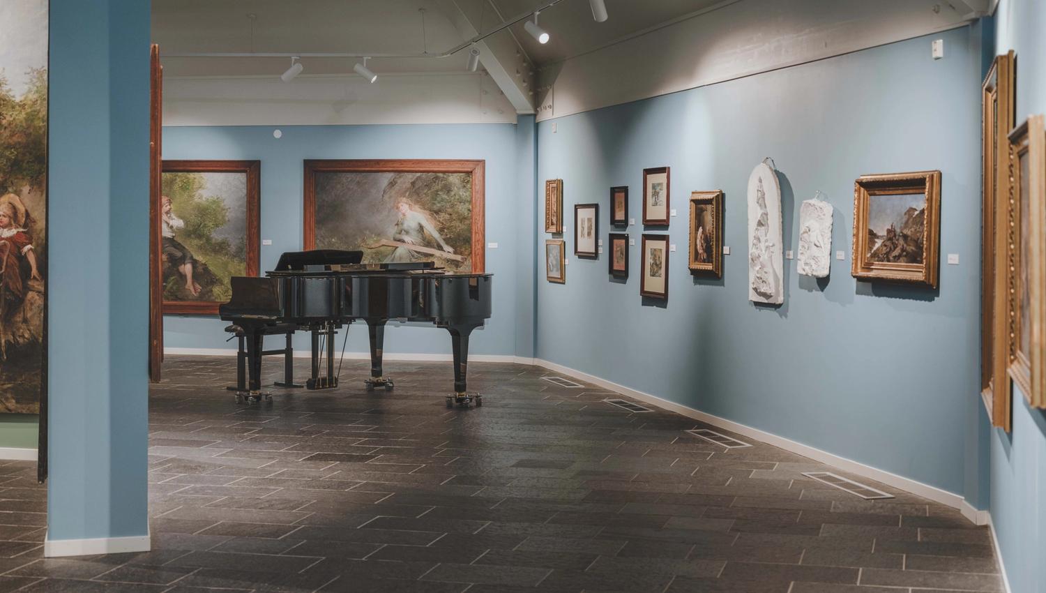 A baby grand piano in front of some of Bergsliens paintings.