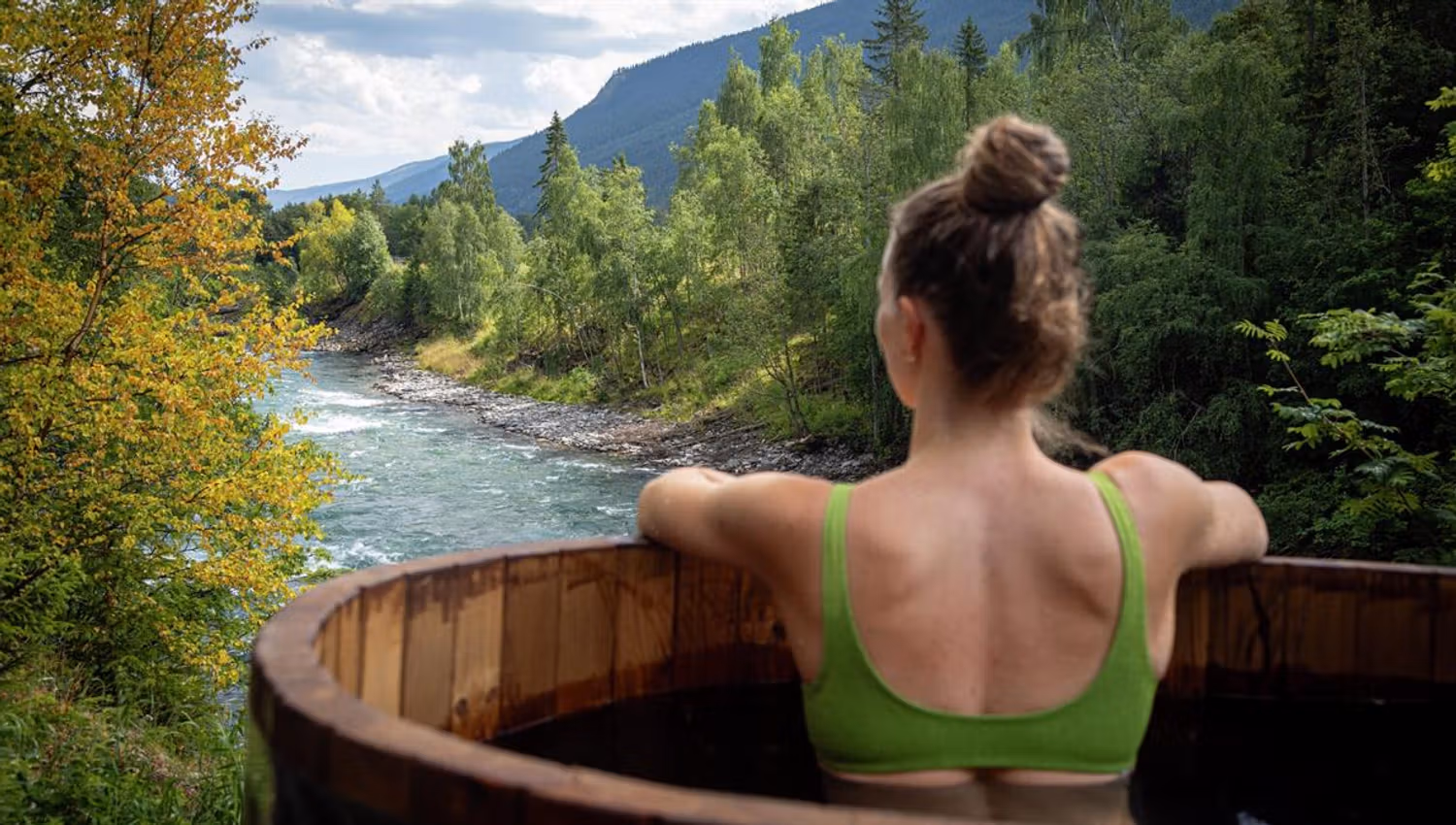 Person sitting in a wooden outdoor hot tub beside a river, with forest and mountains in the background.