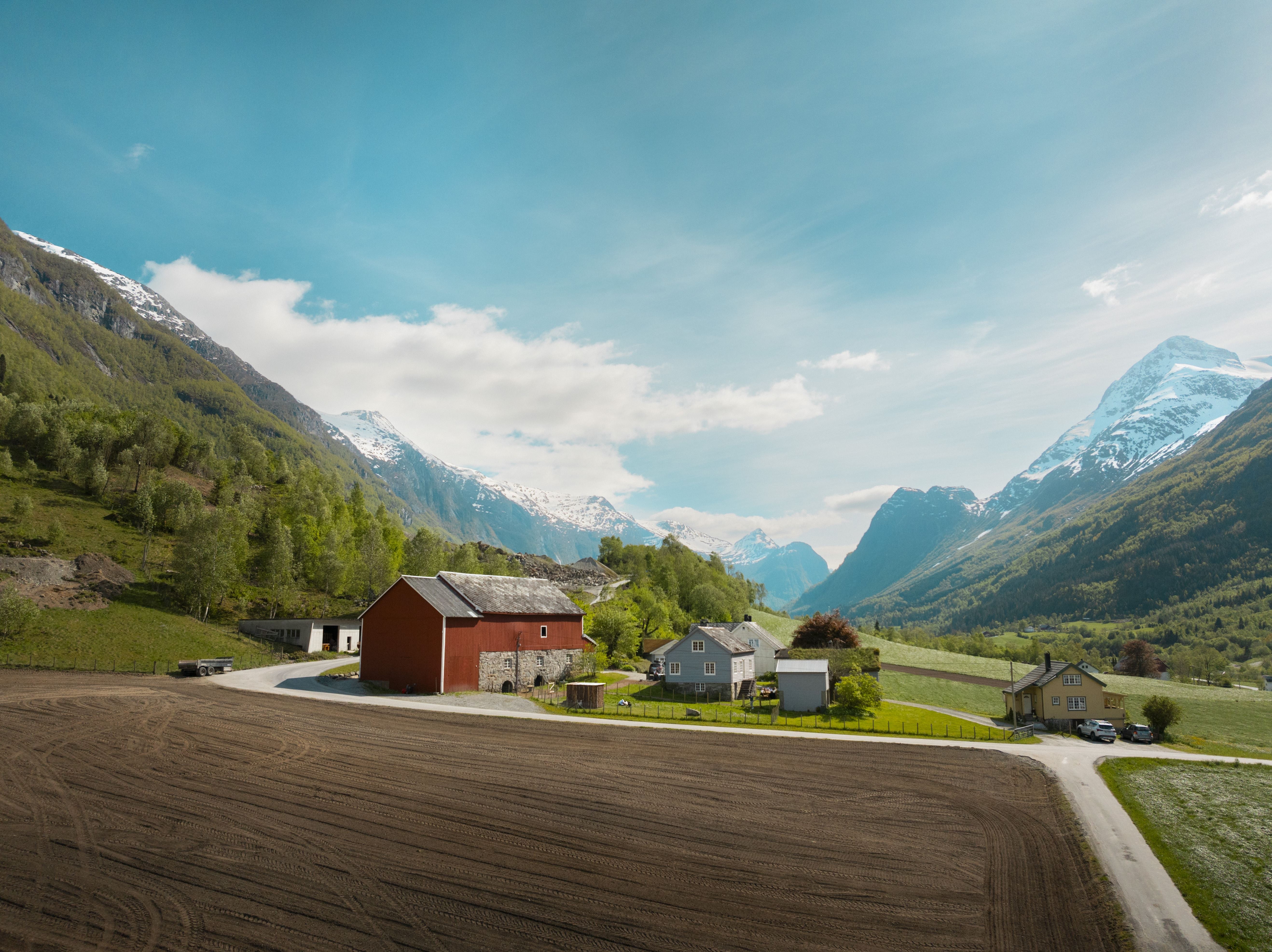 Rural farm buildings beside a plowed field, with green hills and snowcapped mountains under a bright blue sky.