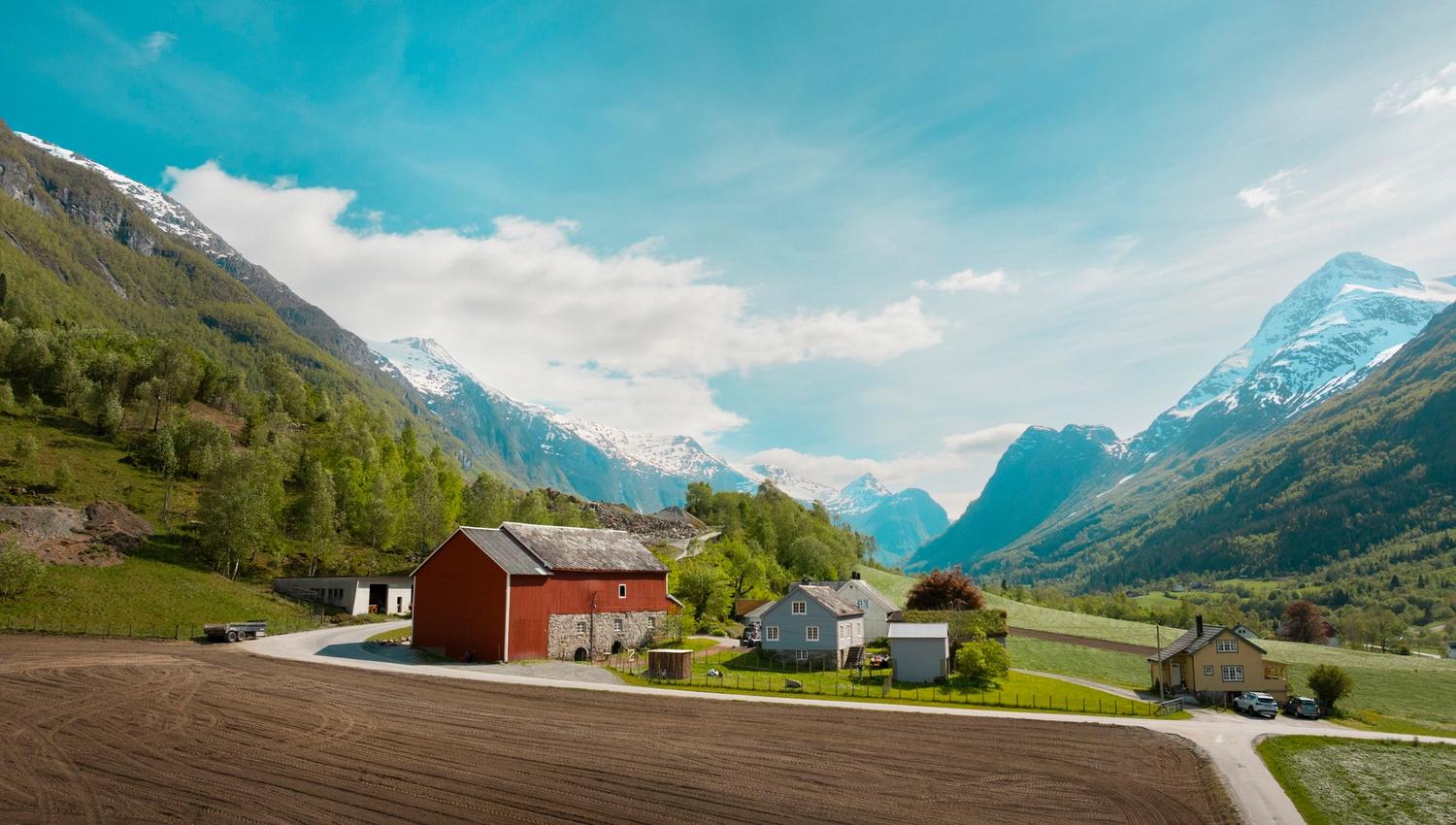 Rural farm buildings beside a plowed field, with green hills and snowcapped mountains under a bright blue sky.