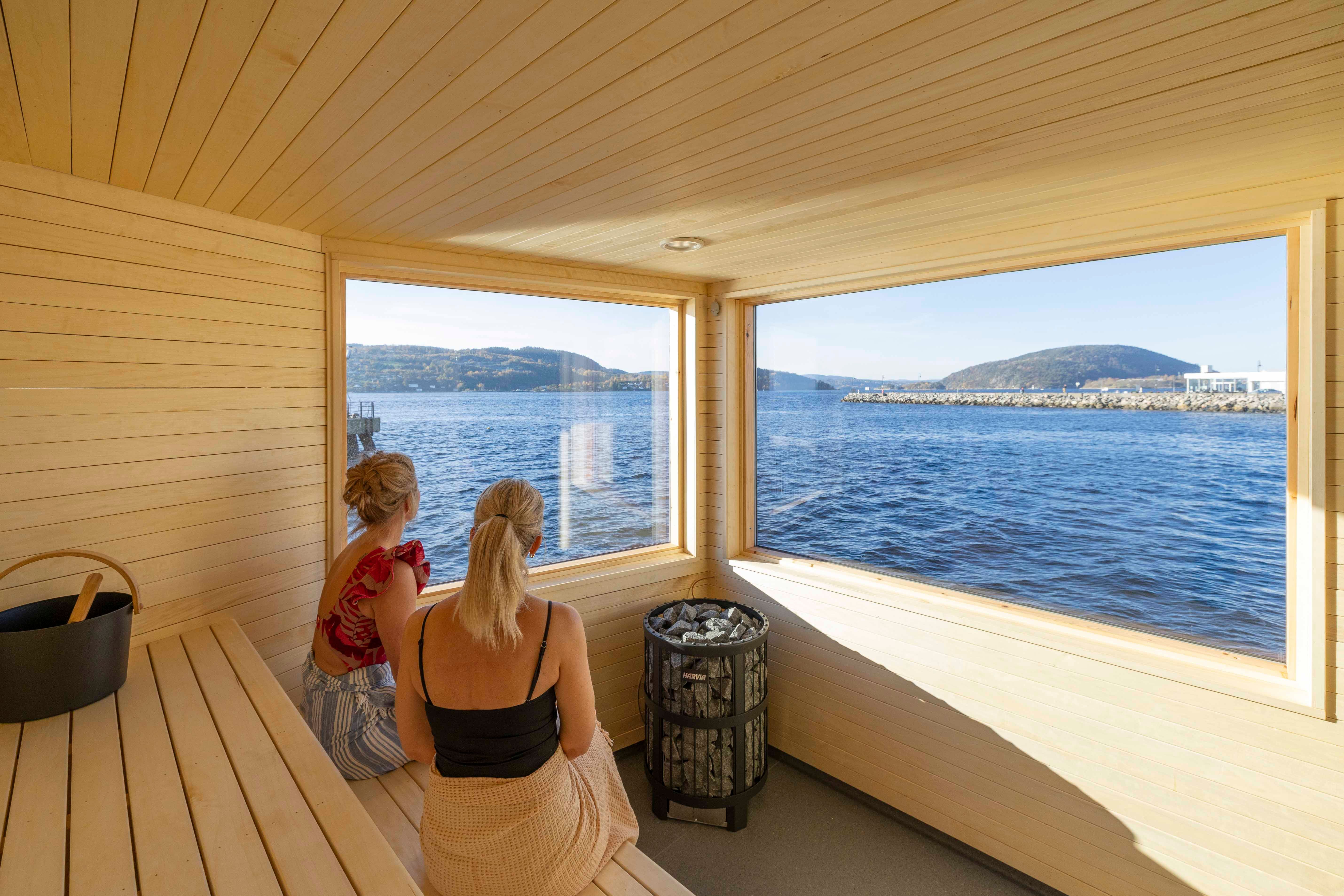 To damer som sitter i badstua med fjordutstikt