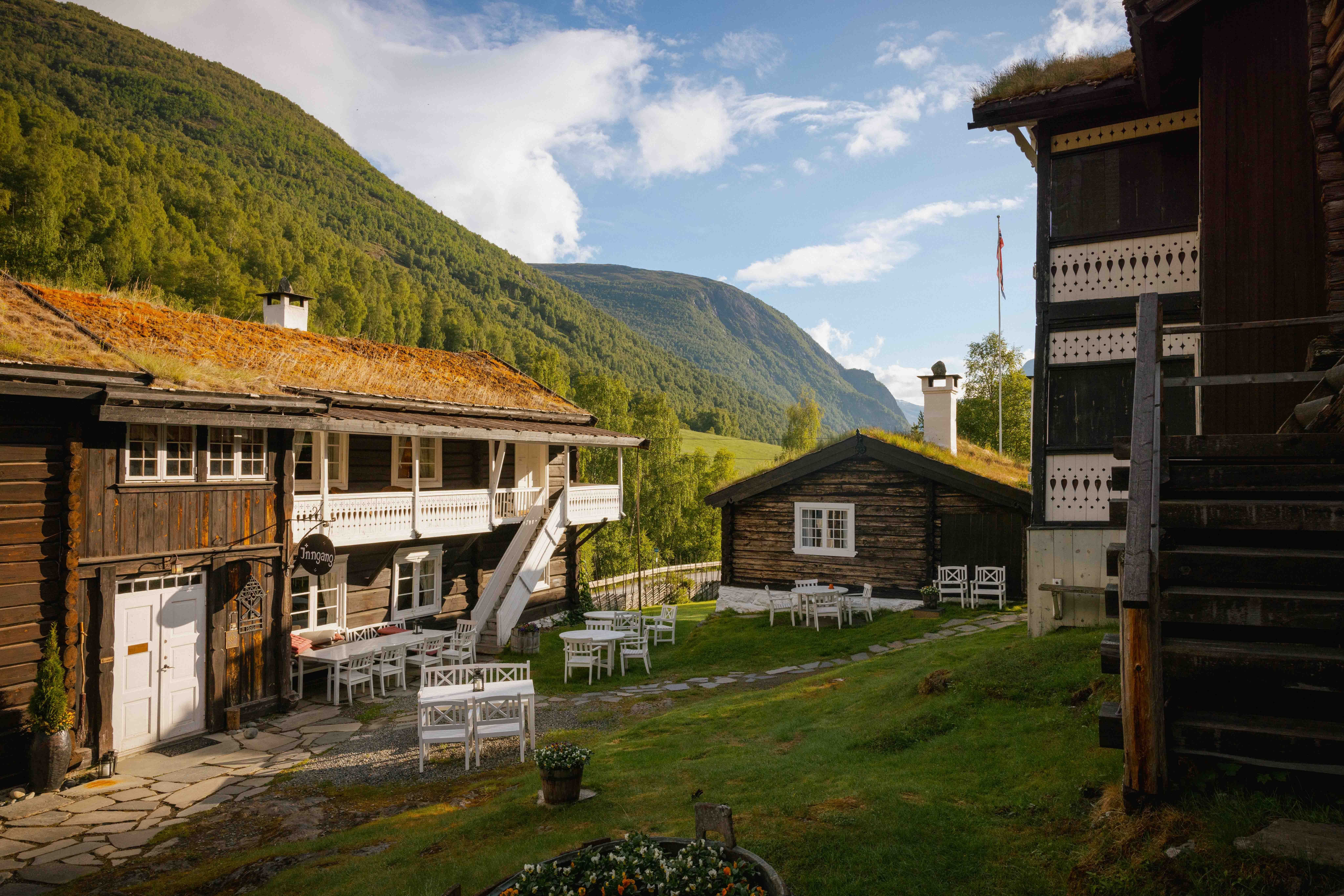 Tun med gamle trehus og kvite utemøblar på grønt gras, med skogkledde fjell og blå himmel i bakgrunnen..