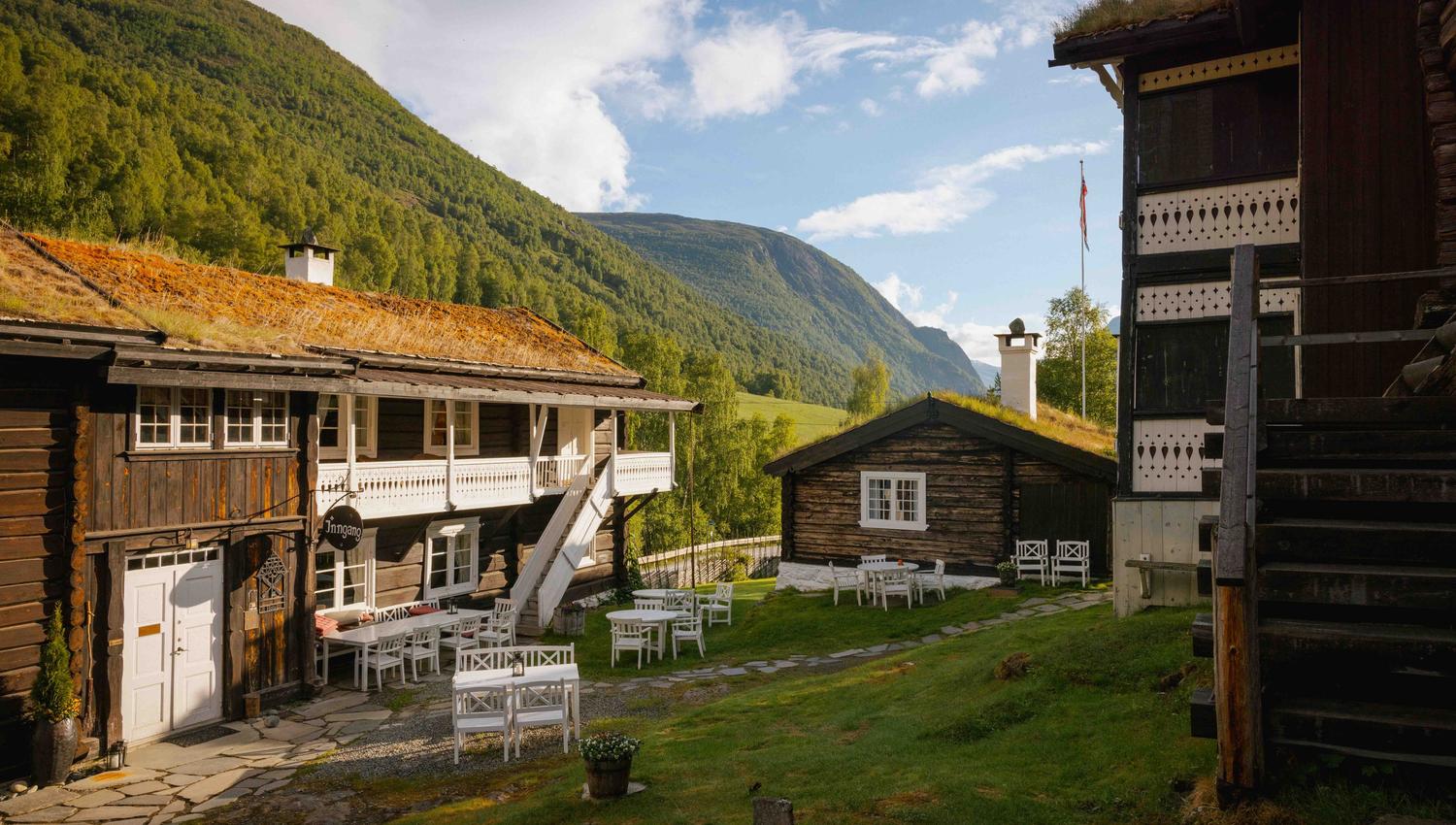 Tun med gamle trehus og kvite utemøblar på grønt gras, med skogkledde fjell og blå himmel i bakgrunnen..