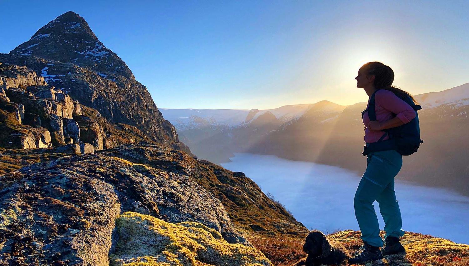 A hiker with a backpack stands on a rocky ridge at sunrise, overlooking a misty lake and mountains..