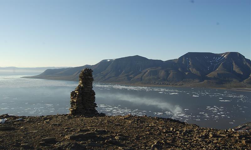 A cairn on top of a mountain with a fjord, a mountain and a clear blue sky in the background