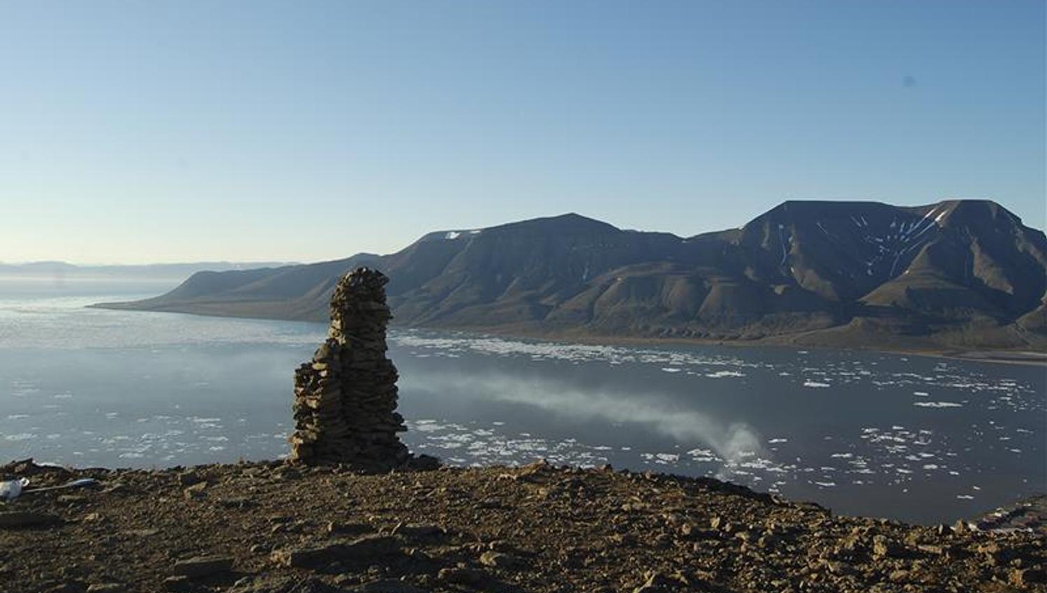A cairn on top of a mountain with a fjord, a mountain and a clear blue sky in the background