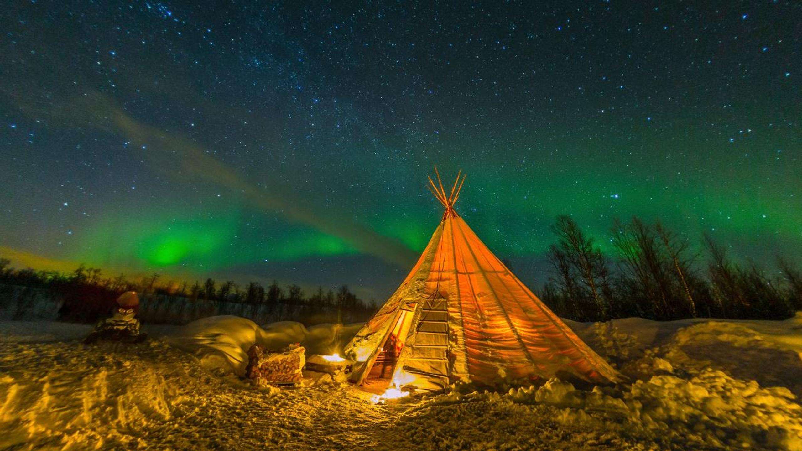 A lavvu in a winter landscape beneath green Northern Lights and a starry sky.