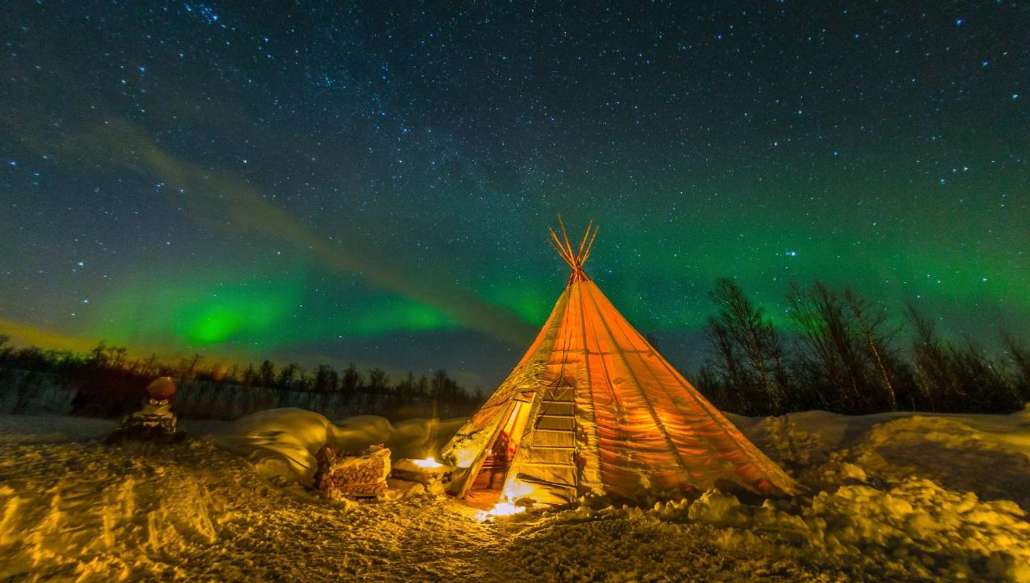 A lavvu in a winter landscape beneath green Northern Lights and a starry sky.