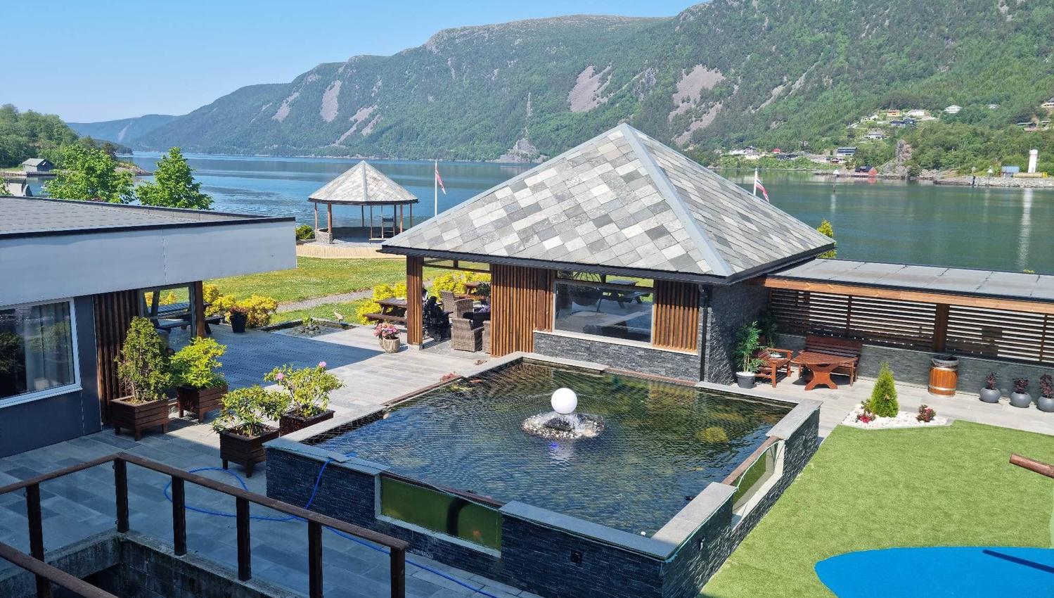 Moderne bygg ved ein fjord med fjell i bakgrunnen, hageanlegg og rektangulært basseng med fontene..