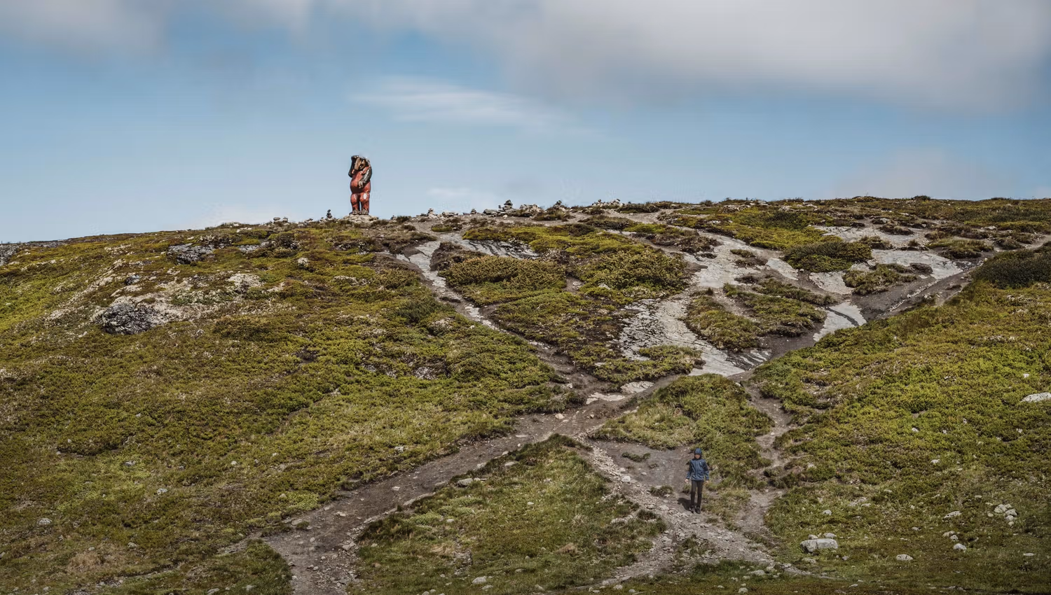 A large wooden troll sitting on top of a hill. A child is standing on a path below the troll.