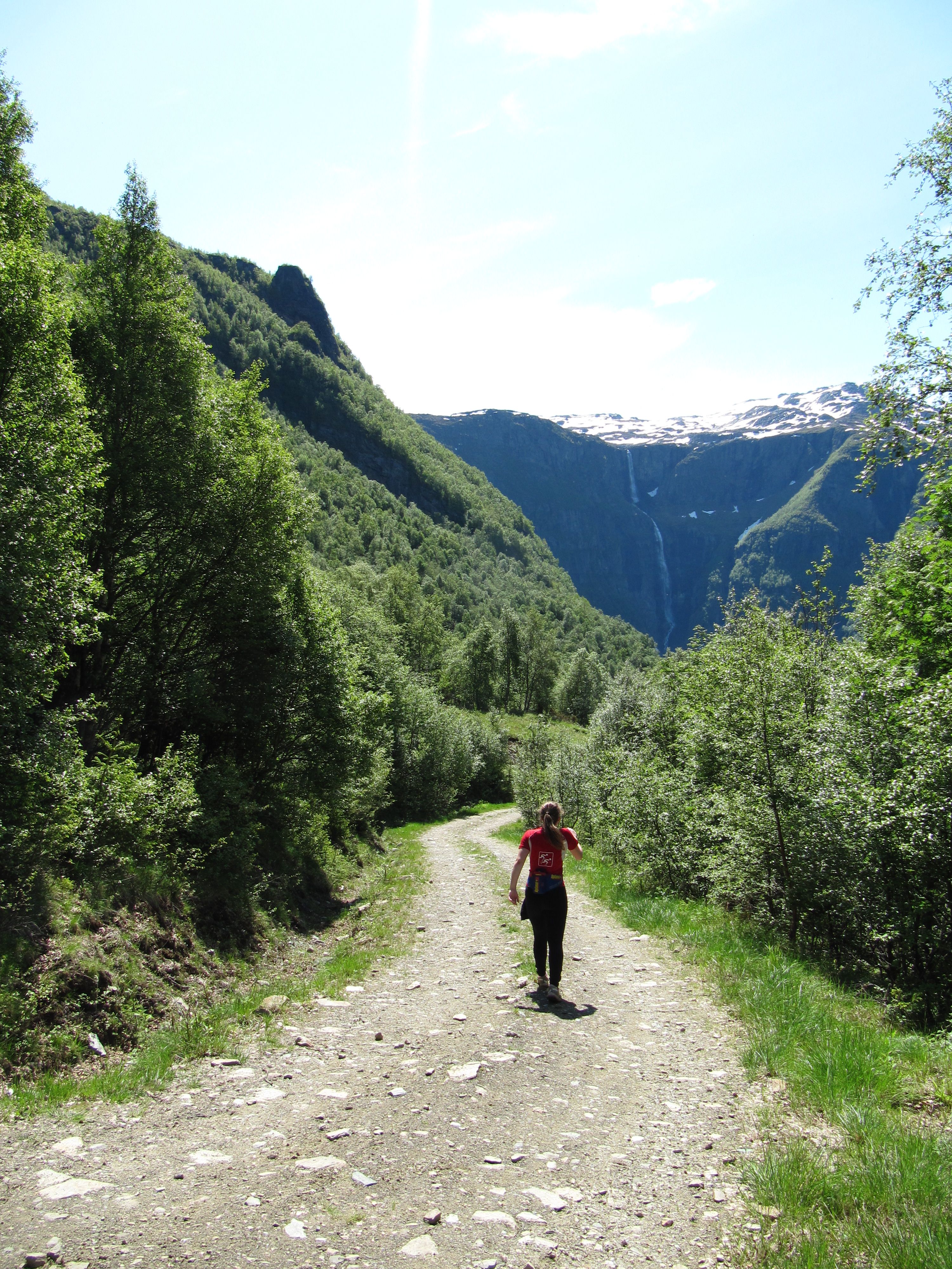 Hiker walking along the gravel road toward the Monk’s Steps with a waterfall and glaciers in the background.