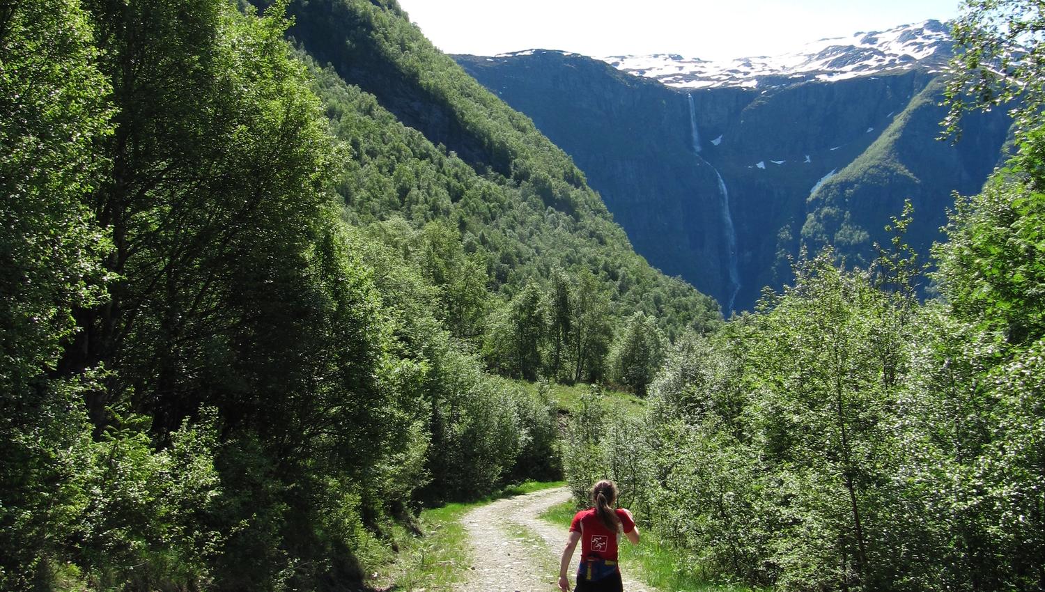 Hiker walking along the gravel road toward the Monk’s Steps with a waterfall and glaciers in the background.