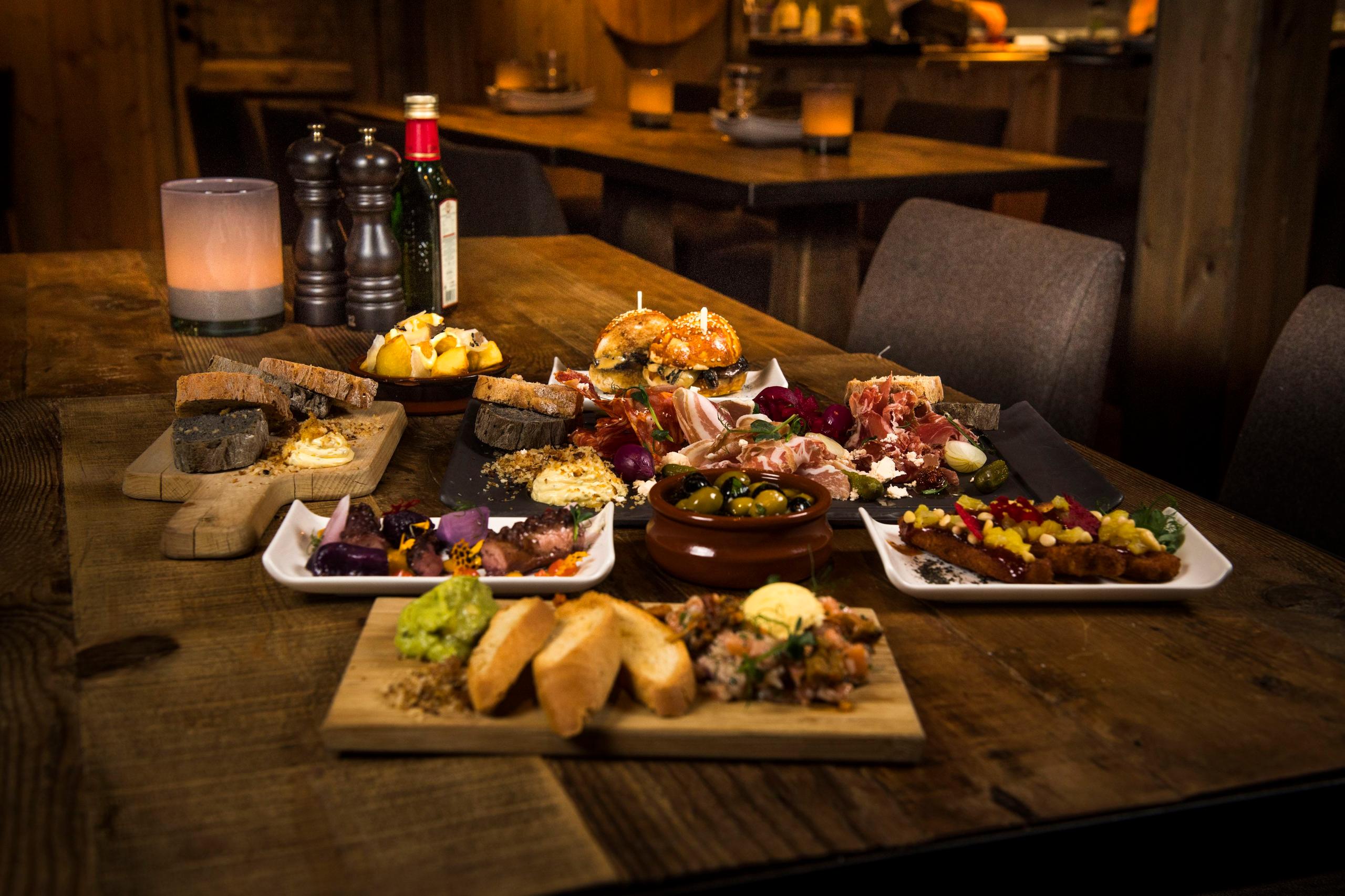 Rustikt trebord dekket med tapas, spekemat, oliven, brød og småretter i en dempet, varm restaurant..