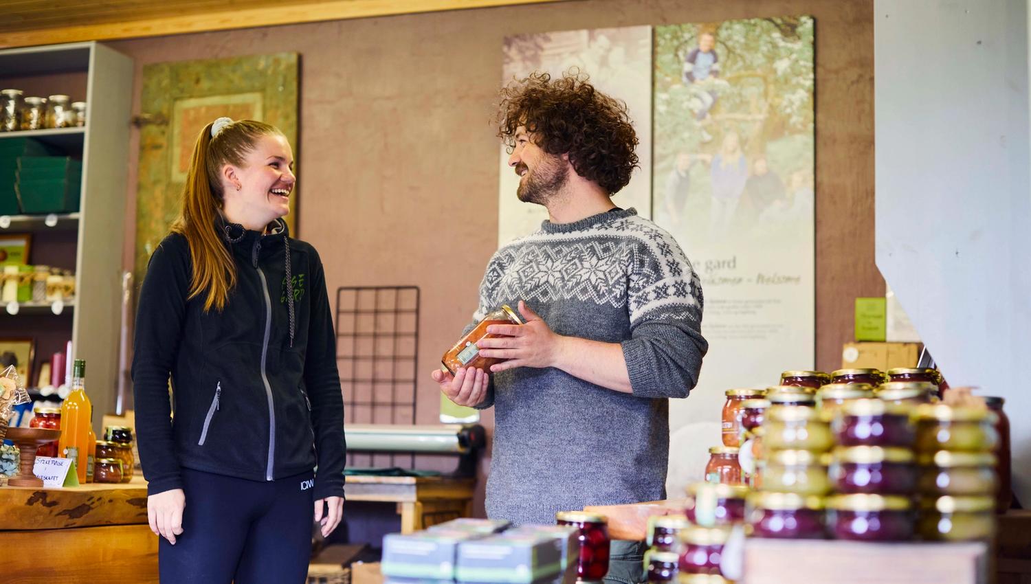 Zwei Mitarbeitende unterhalten sich lachend im Hofladen von Syse Farm, umgeben von regionalen Produkten und hausgemachter Marmelade.