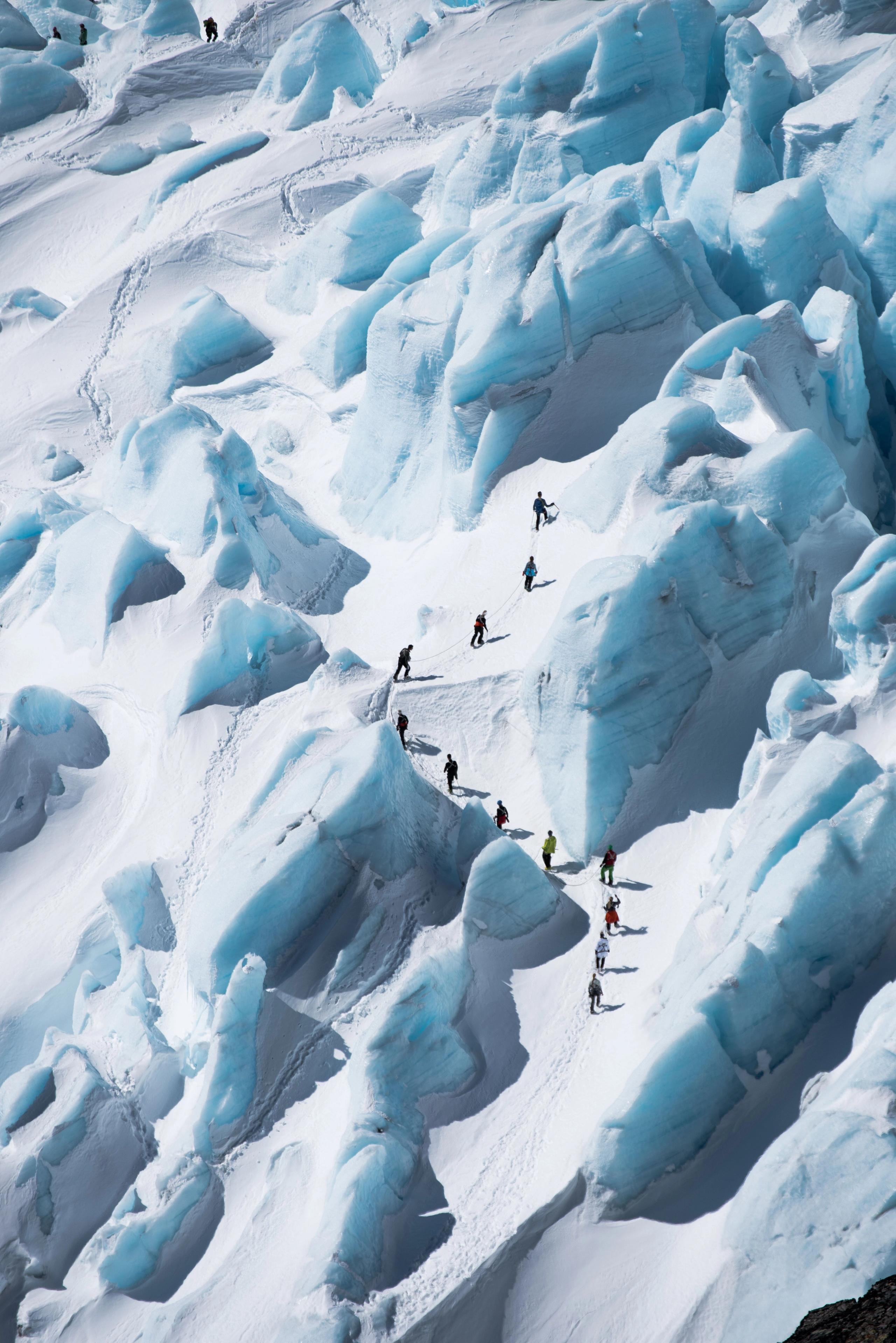 A line of mountaineers traverses a snowy glacier amid jagged blue ice seracs and deep crevasses in daylight..