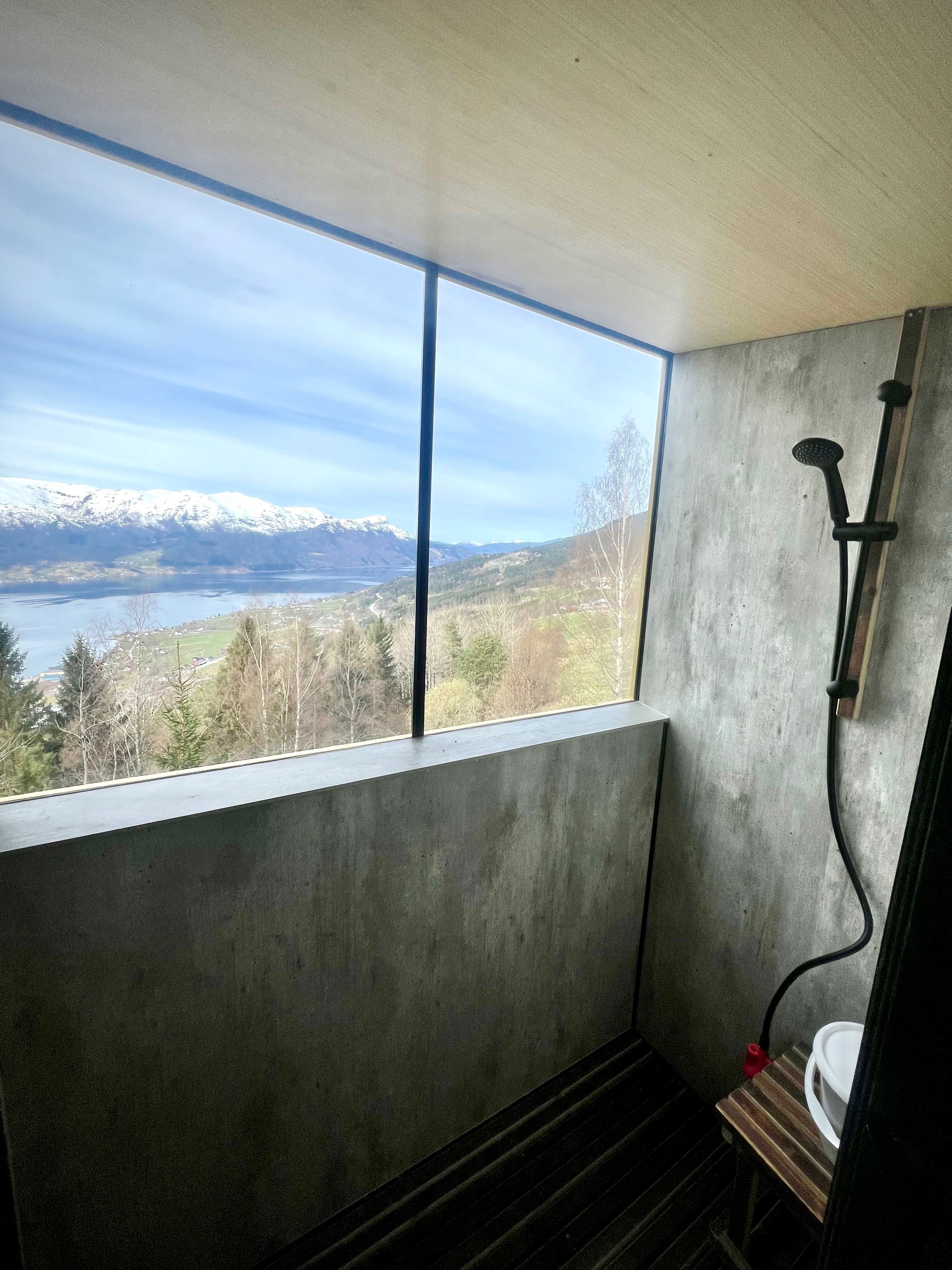 Concrete shower stall with black showerhead beside a large window overlooking a lake, trees, and snowy mountains..