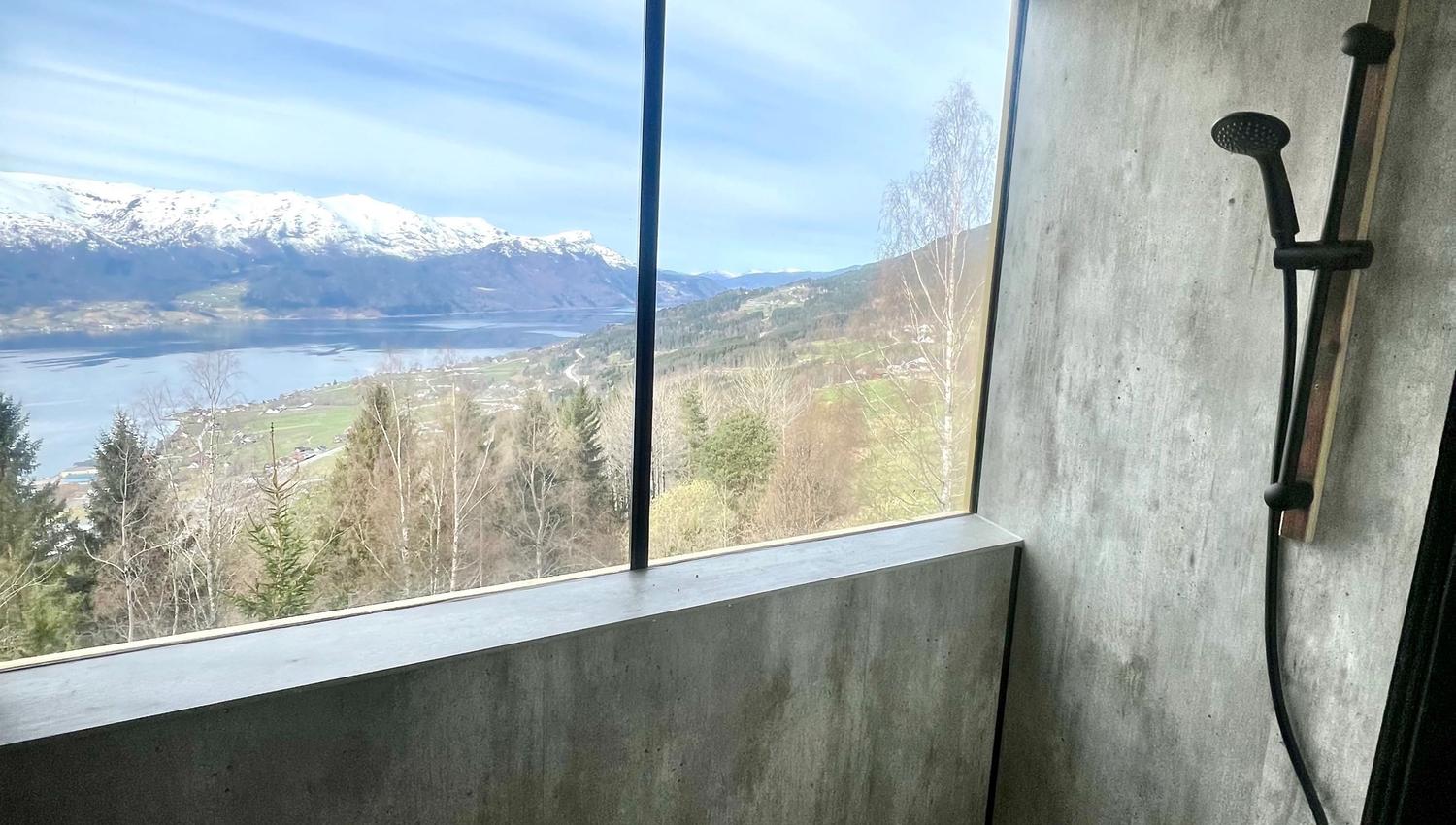 Concrete shower stall with black showerhead beside a large window overlooking a lake, trees, and snowy mountains..