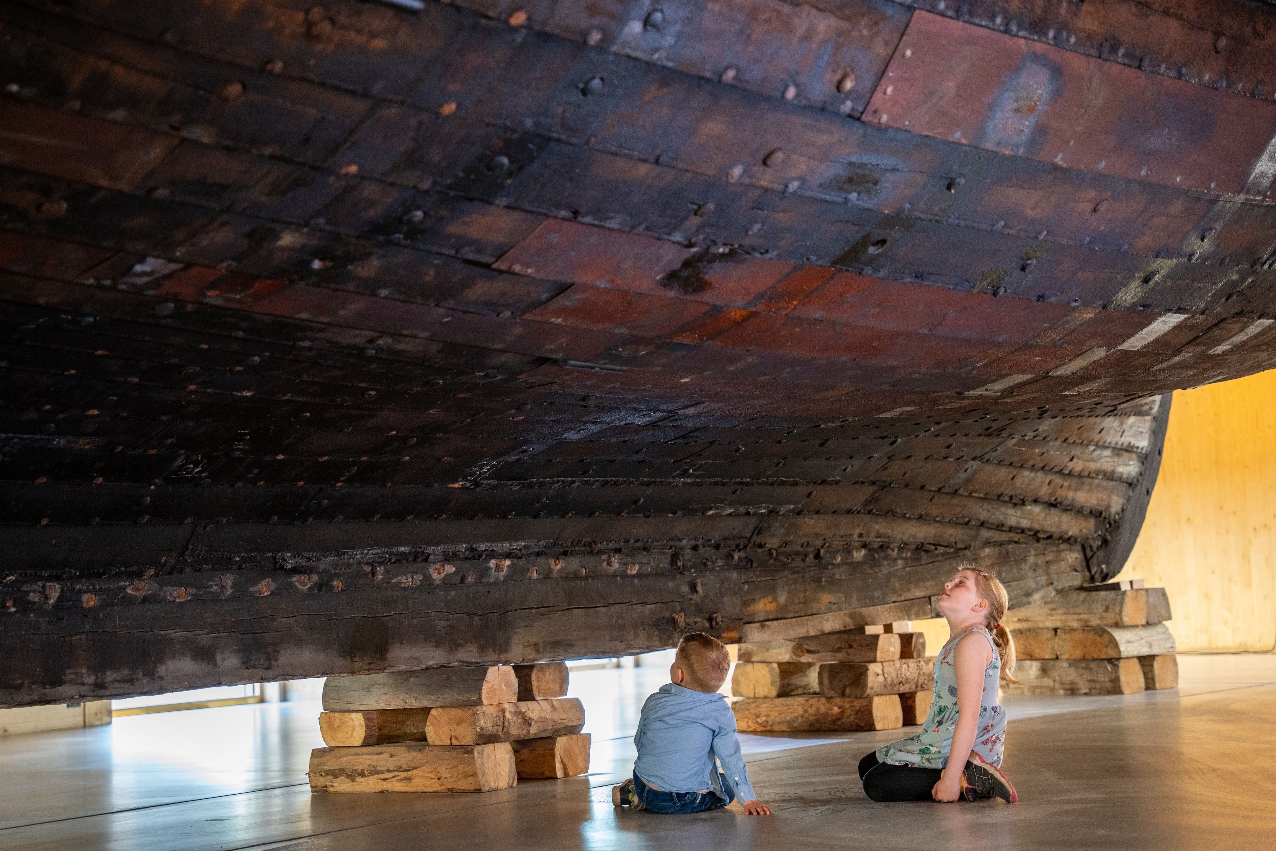 Two children sit on a museum floor beneath a large wooden ship hull supported by stacked timber blocks..