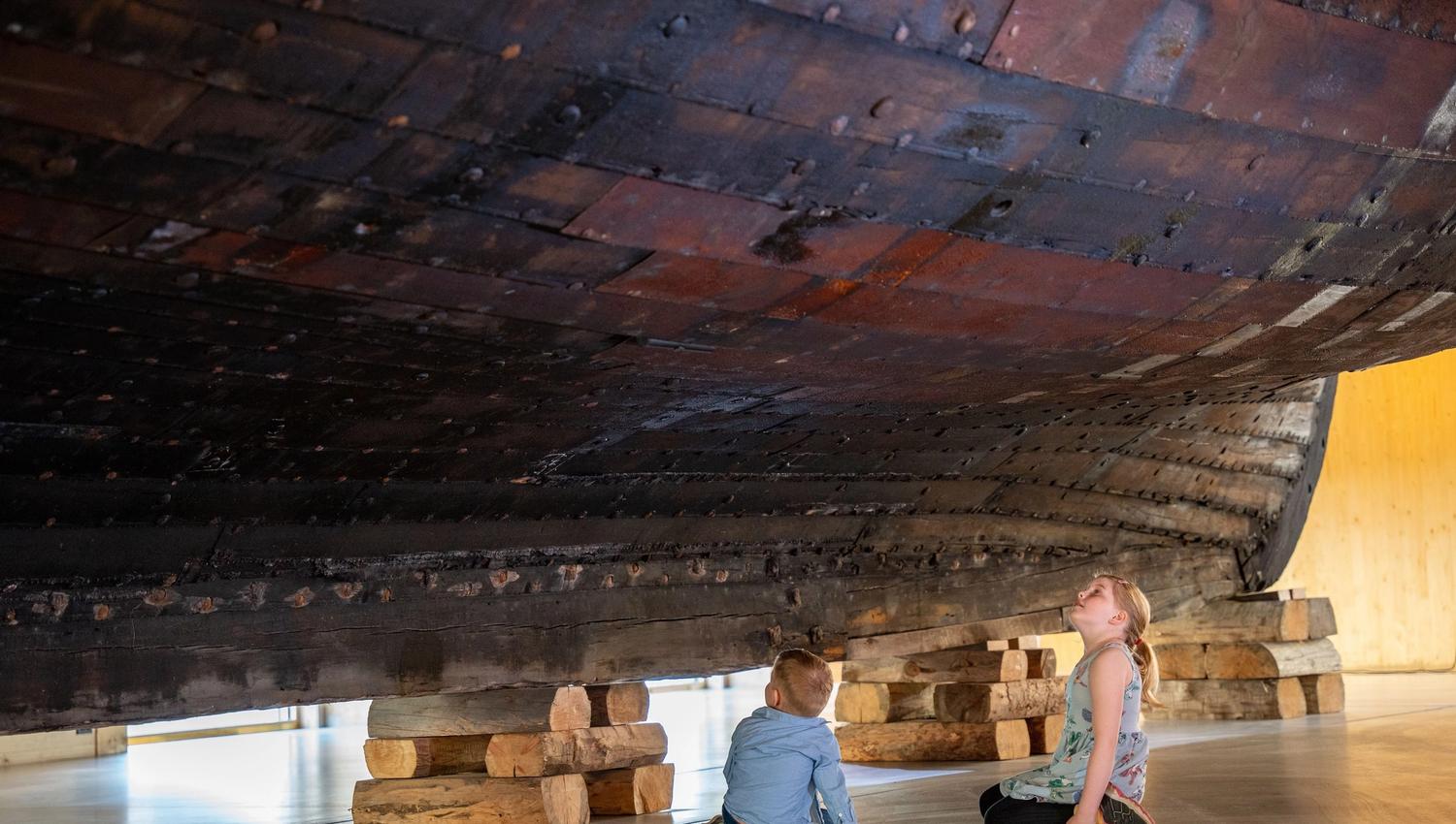 Two children sit on a museum floor beneath a large wooden ship hull supported by stacked timber blocks..